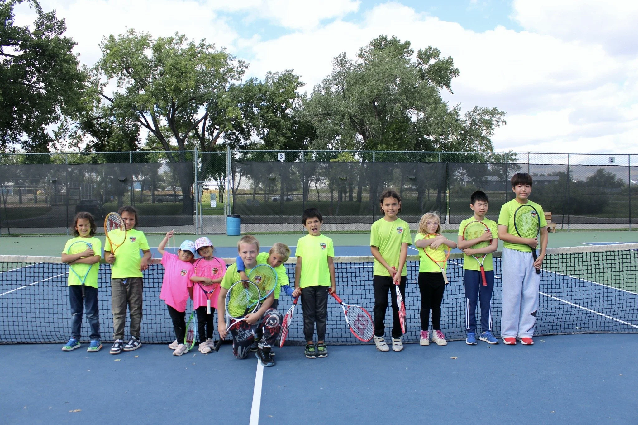 Group of children on a tennis court holding tennis rackets, some smiling and posing for the camera, with a background of trees and sky.