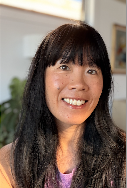 A woman with long black hair and bangs, smiling indoors with a plant in the background.
