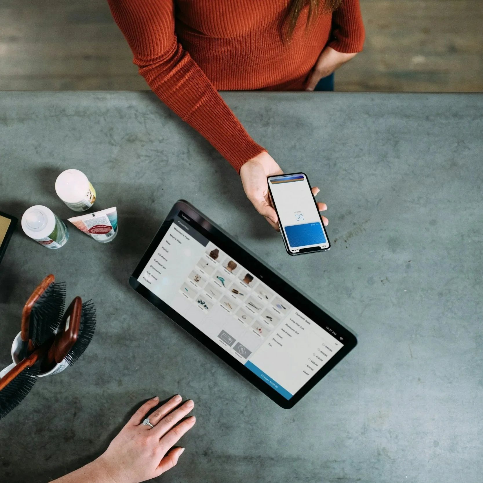 Overhead view of a person in an orange sweater using a smartphone, with a tablet displaying an online shopping site on a concrete countertop, along with various bottles, grooming tools, and hairbrushes.