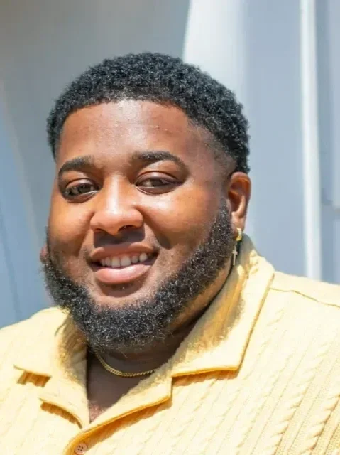 A smiling man with curly hair and a beard, wearing a yellow shirt and a necklace, standing indoors with part of a window or door frame visible in the background.