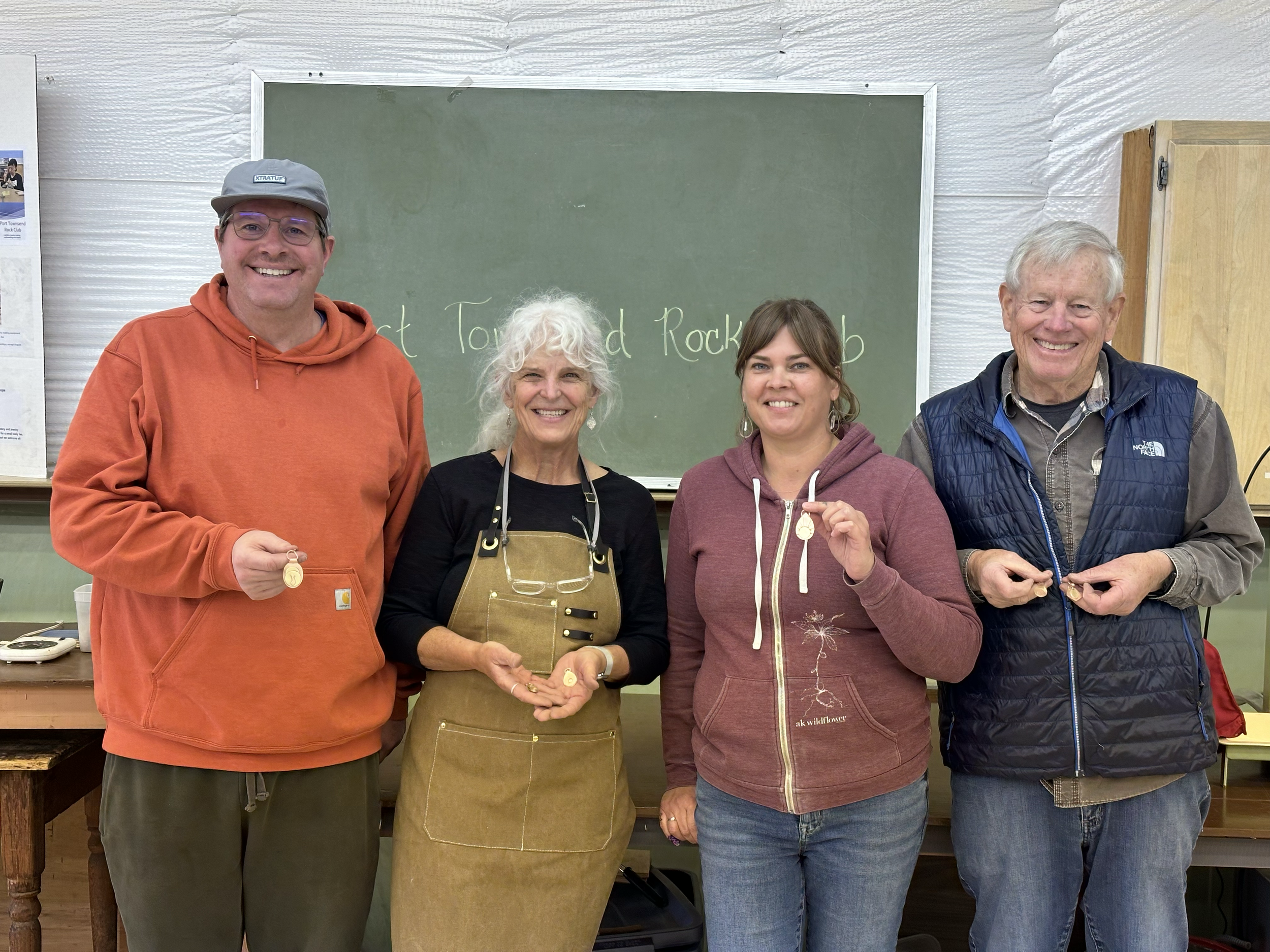 Four people standing in front of a chalkboard, smiling and holding small awards or medallions. The chalkboard has some writing that is partially visible, mentioning 'Rock...' and 'Club'.