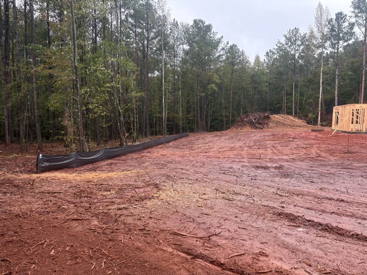 A cleared construction site with dirt ground, a black landscape fabric or silt fence along the edge, and a partially built wooden structure in the background amidst trees.