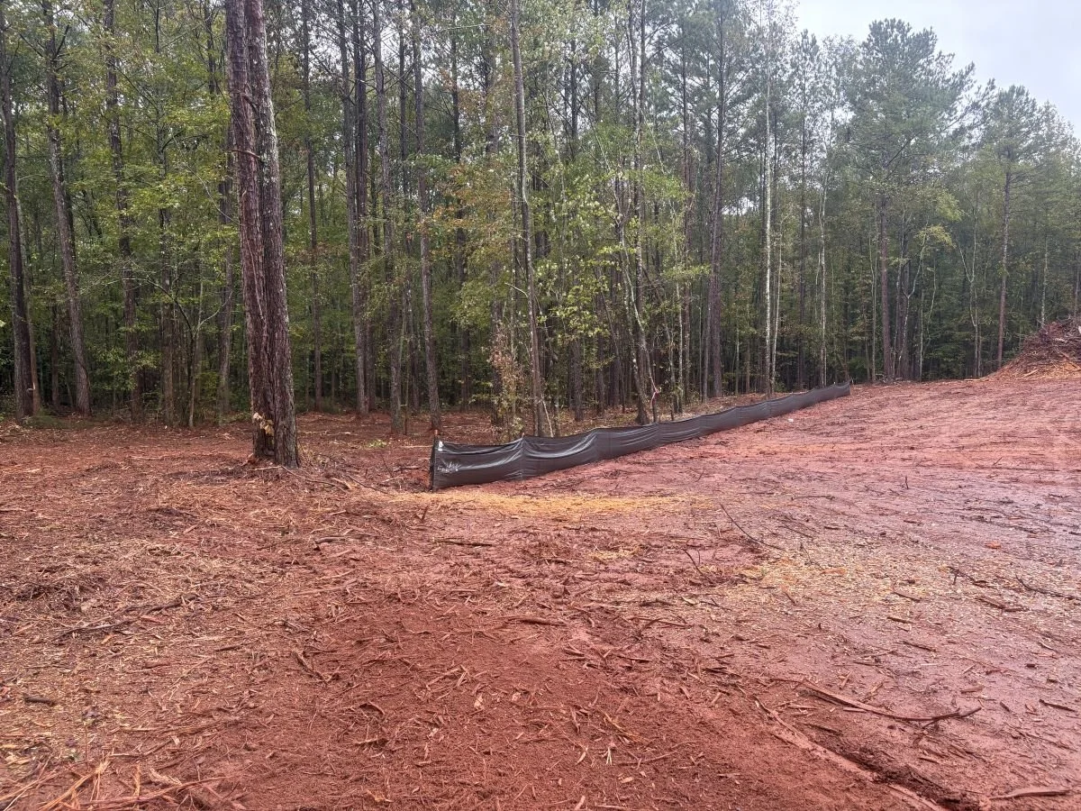 A wooded area with a cleared patch of reddish soil and a black erosion control barrier along the edge.