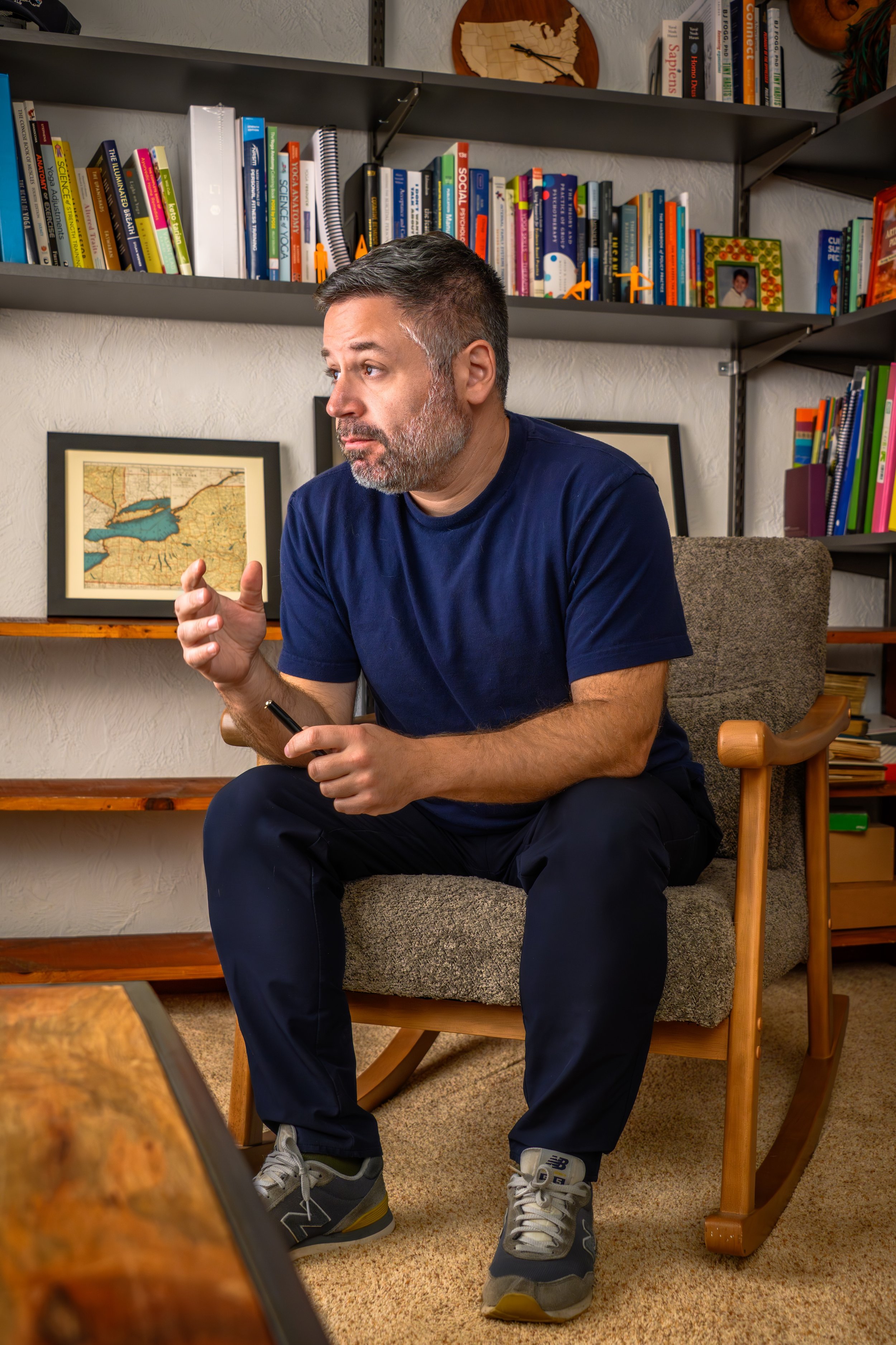 A man with short gray hair and beard, wearing a navy blue t-shirt and dark pants, sitting on a wooden chair with a cushioned seat in a room full of bookshelves. He is holding a pen and appears to be in a thoughtful or explaining pose.