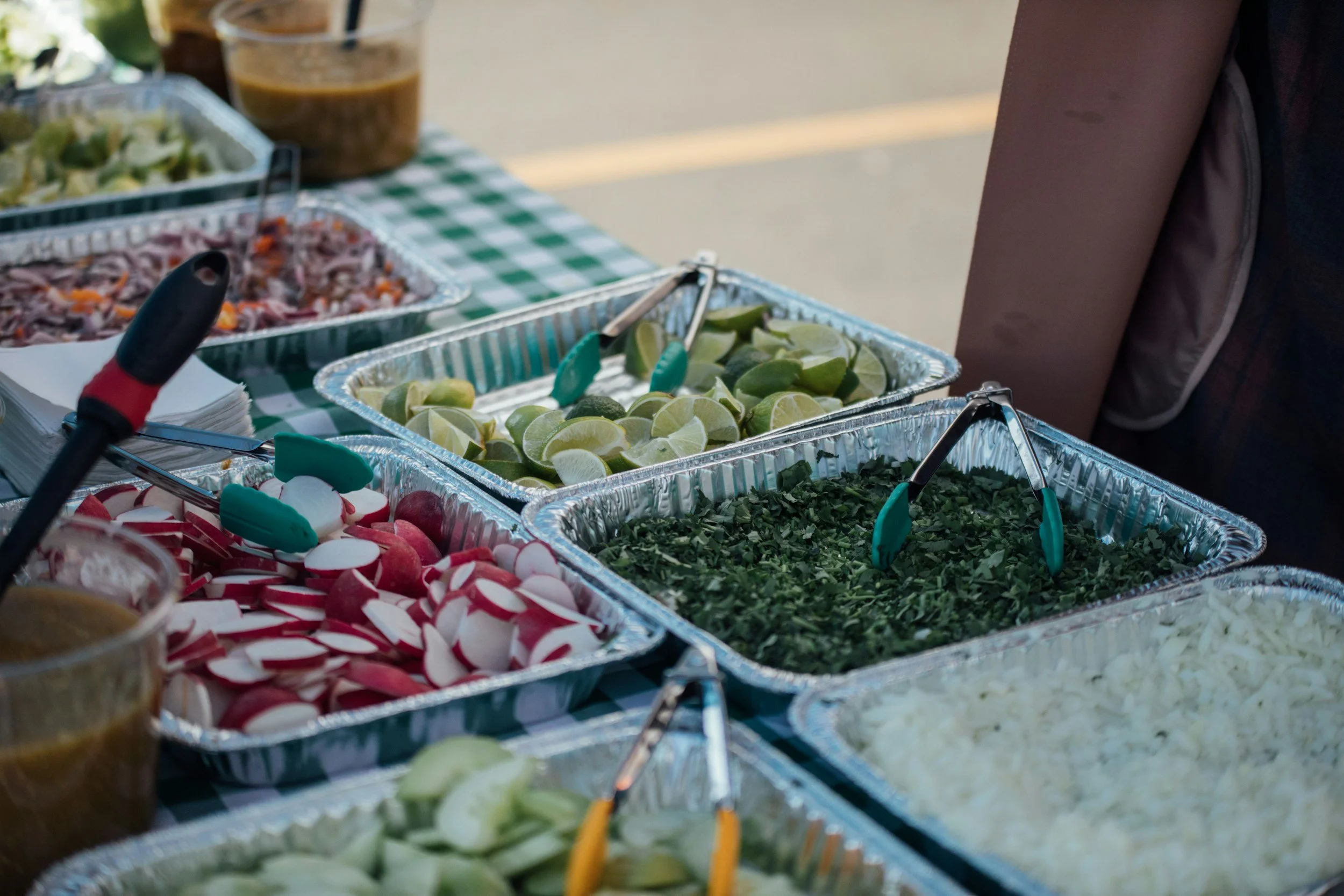 A buffet table with various chopped vegetables, lime wedges, and condiments in aluminum trays, with tongs for serving.