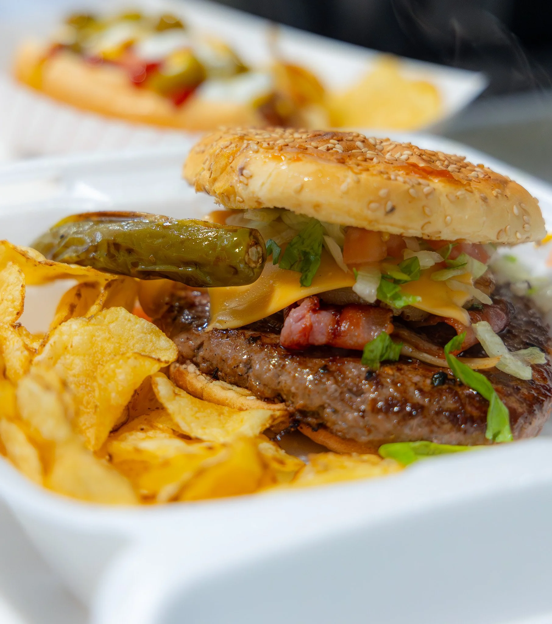 Close-up of a cheeseburger with lettuce, tomato, pickles, cheddar cheese, bacon, and sesame seed bun, served with potato chips.