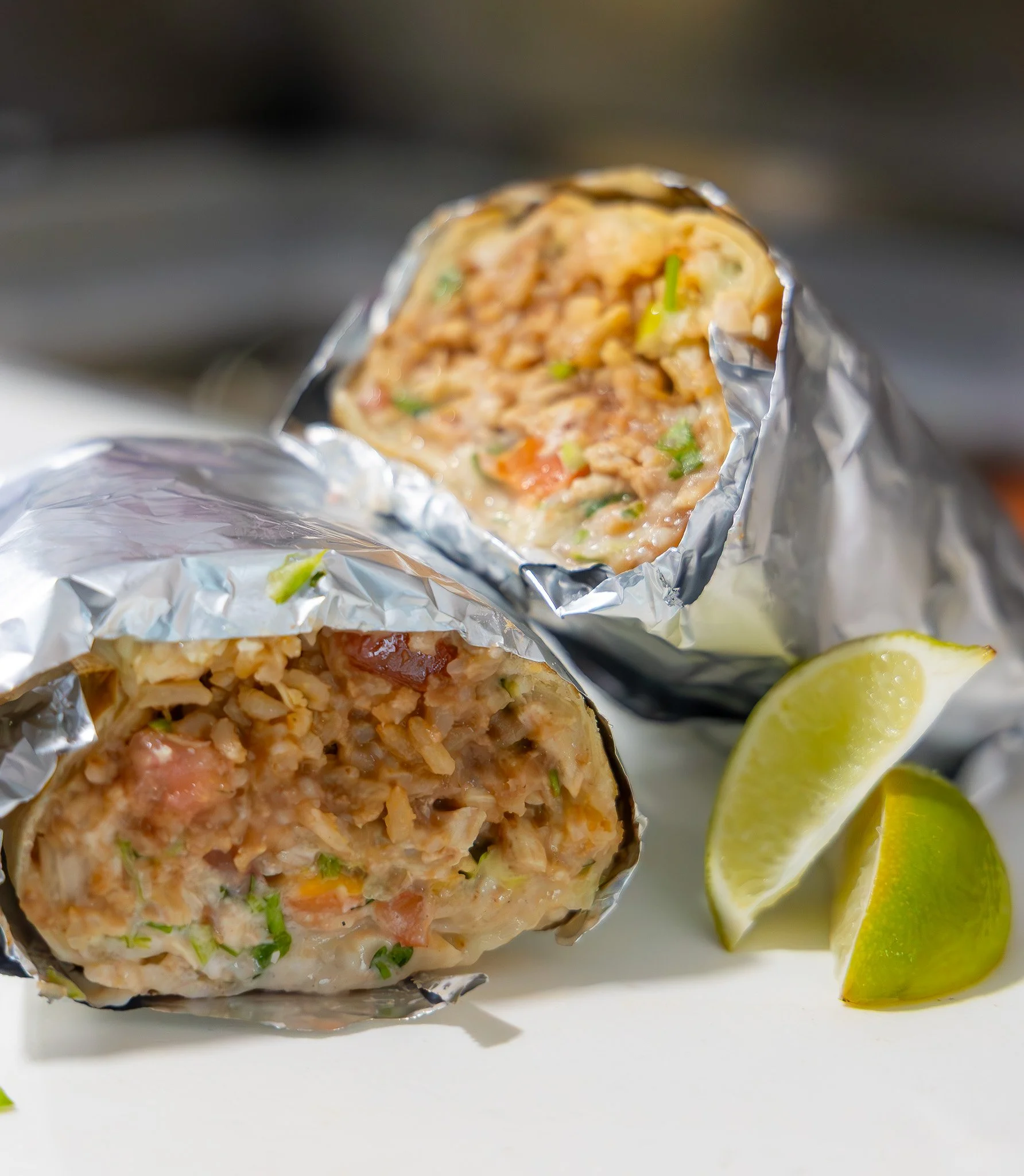 Two burritos wrapped in foil, cut in half showing rice, beans, vegetables, and meat filling inside, with lime wedges beside them.