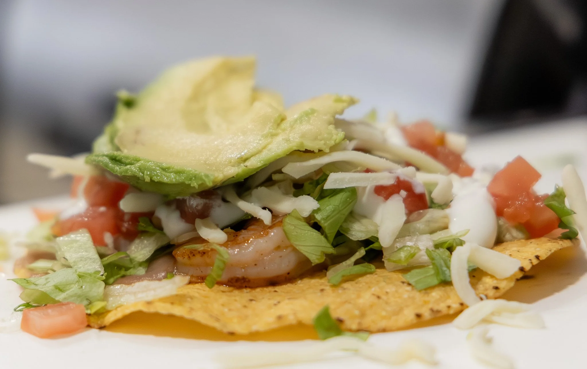 Close-up of a taco with shrimp, lettuce, tomato, shredded cheese, avocado slices, and sour cream on a white plate.