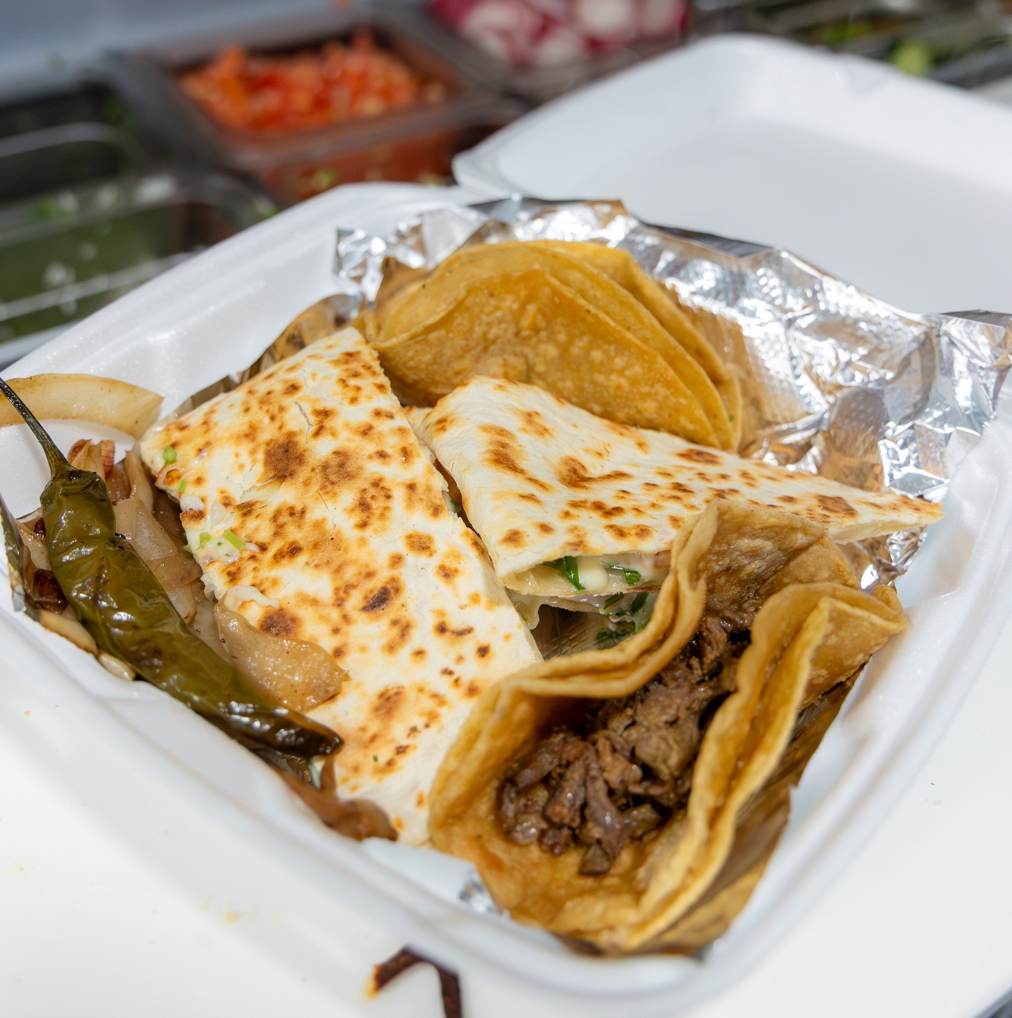 Close-up of a styrofoam takeout container with tacos, a grilled pepper, and a sprinkled cheese quesadilla inside.