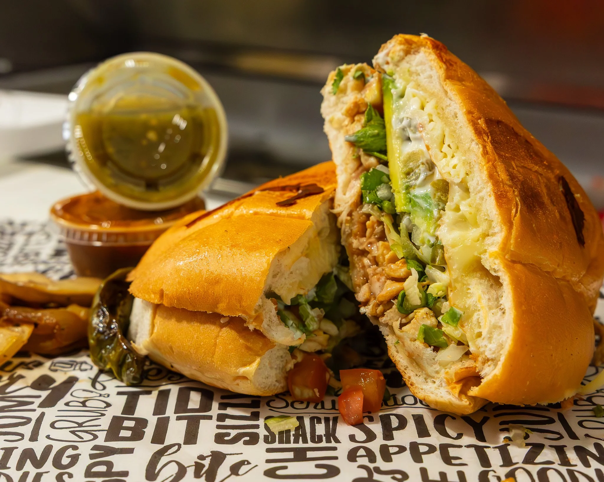 A chicken sandwich cut in half revealing lettuce, avocado, and chicken, served with dipping sauces and caramelized onions on decorative paper.