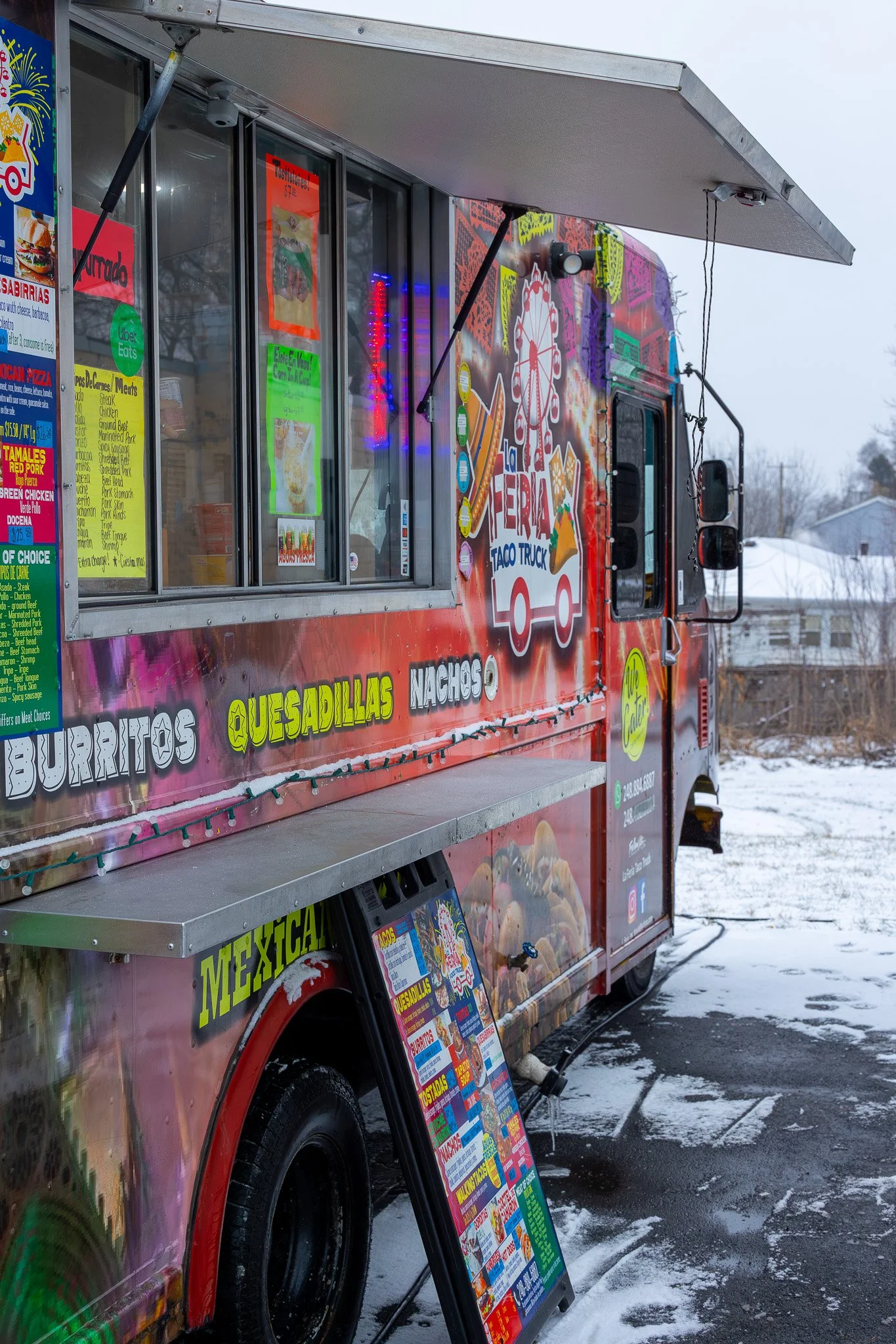 A colorful food truck decorated with neon and winter holiday lights is parked in a snowy outdoor area. The truck advertises Mexican food items like burritos, quesadillas, nachos, and tacos, with a menu displayboard outside.