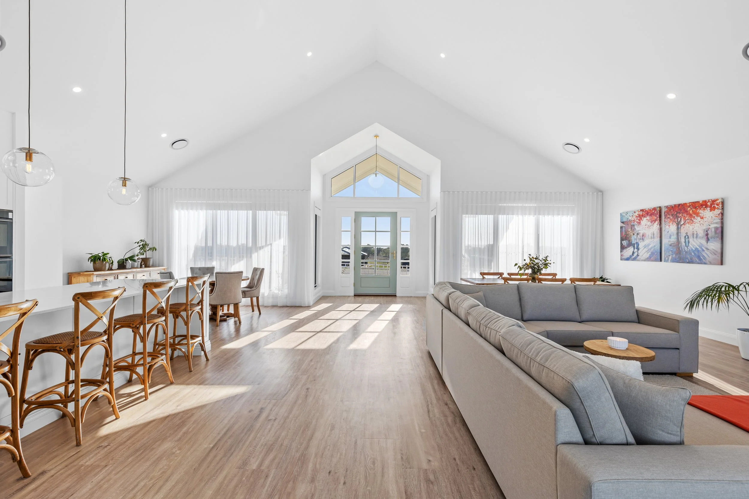Bright, spacious living room with high vaulted ceiling, large window doors, and white walls. Features a light-colored sectional sofa, a wooden coffee table, and framed artwork on the wall. Adjacent to the living area is a dining table with chairs, with large windows and sheer curtains letting in natural light. A few houseplants are also visible.