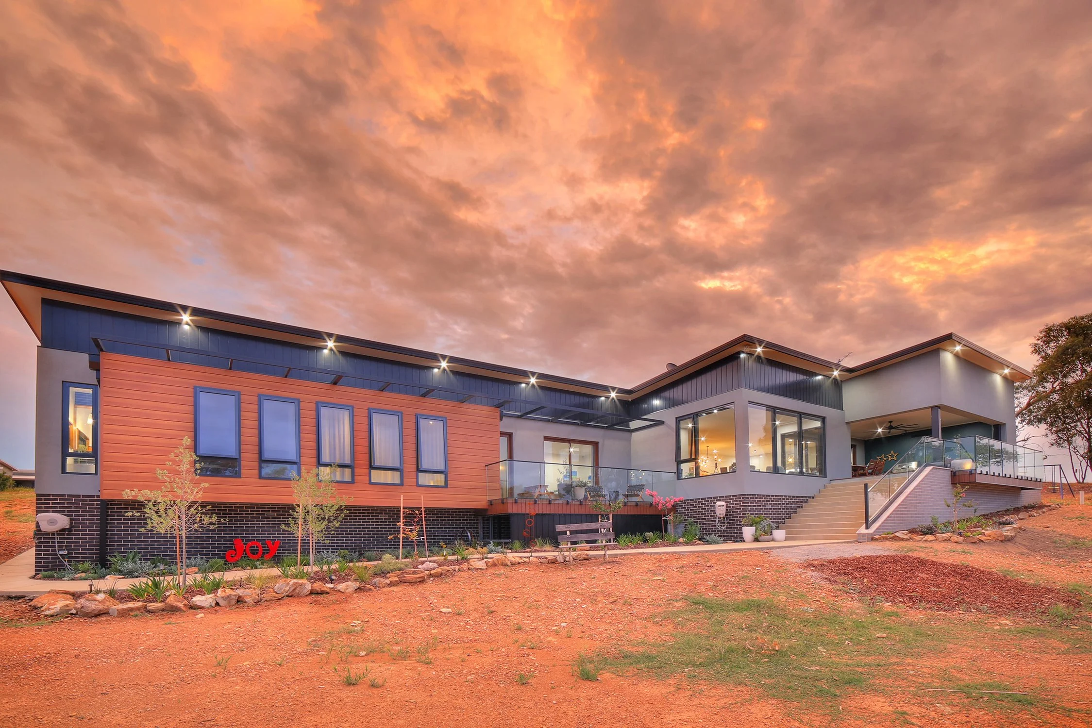 Modern house with large windows and outdoor staircase at sunset, landscaped yard with rocks and plants, word 'Joy' in red on the yard.