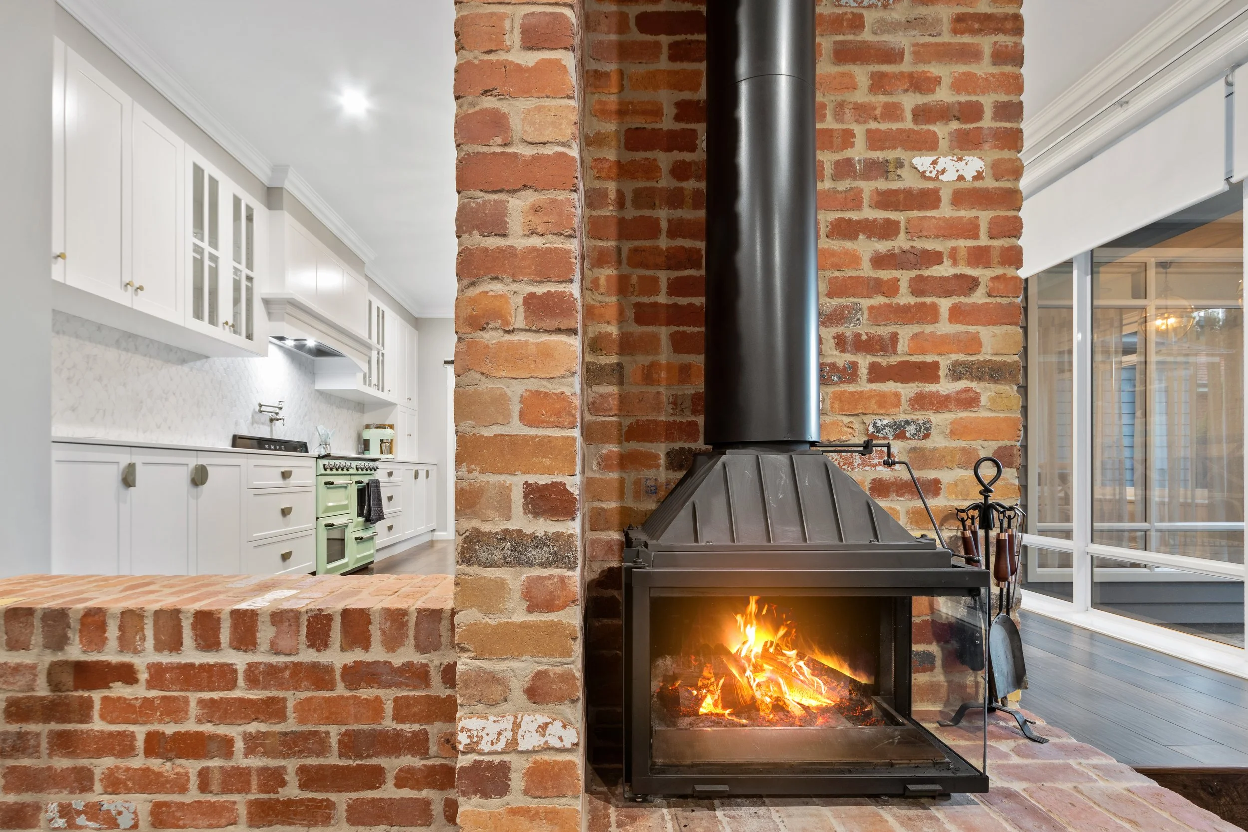 Living room with a brick fireplace with a fire burning, adjacent to a bright white kitchen with white cabinets and a black stove.