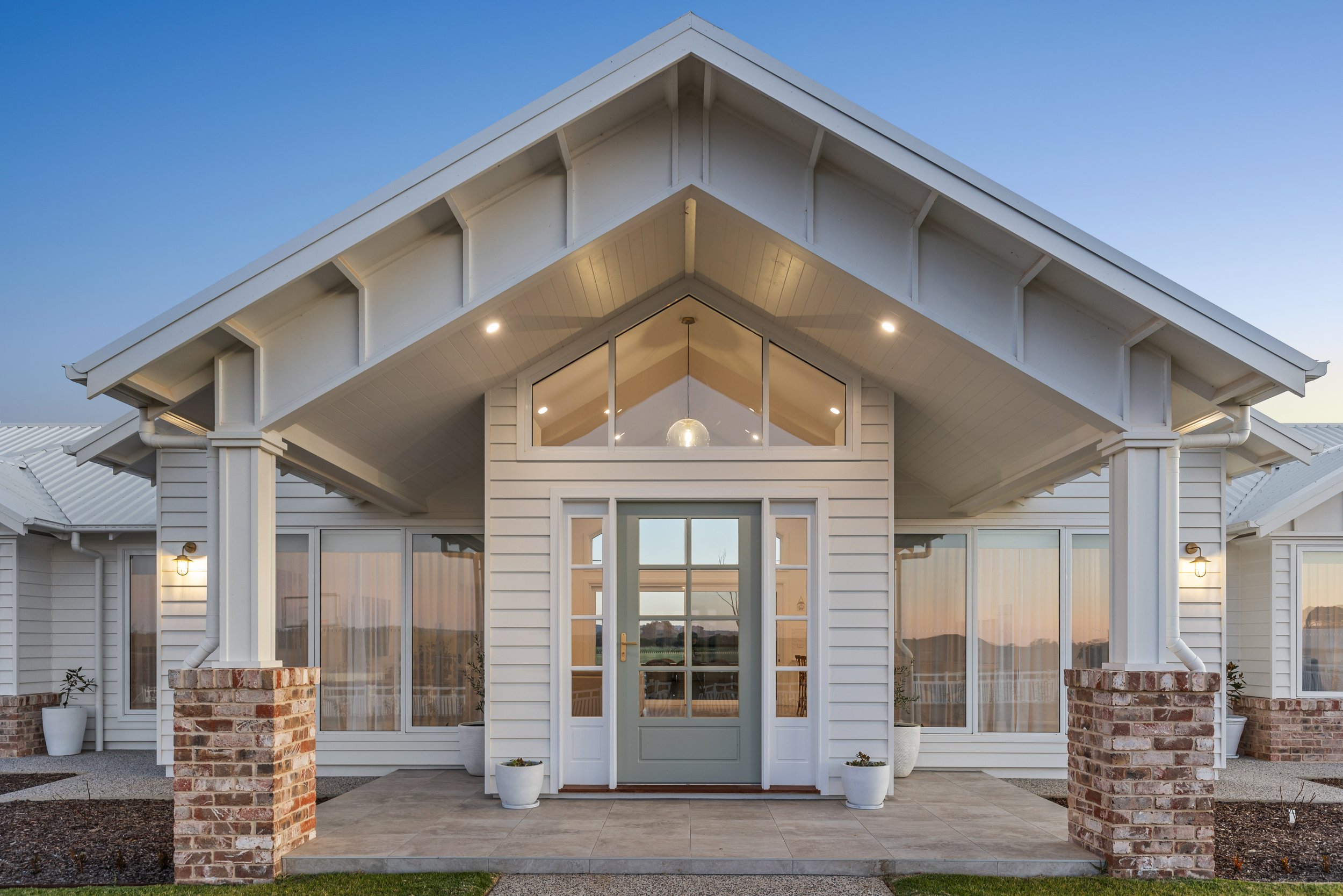 Front view of a modern white house with a gable roof, large glass front door, and windows, surrounded by a landscaped yard with potted plants and brick pillars.