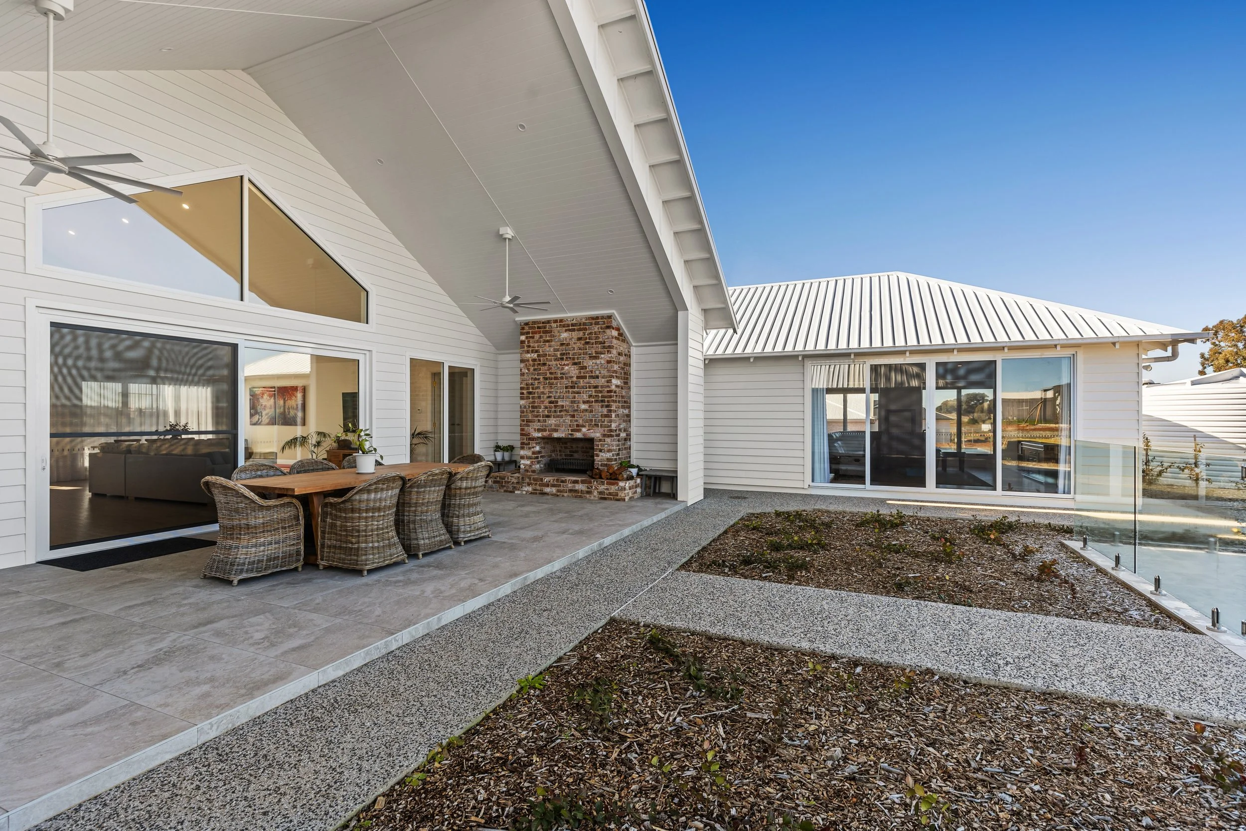 Backyard patio area of a modern house with an outdoor dining table and wicker chairs, a brick fireplace, sliding glass doors, and a gravel garden with a concrete path under a clear blue sky.