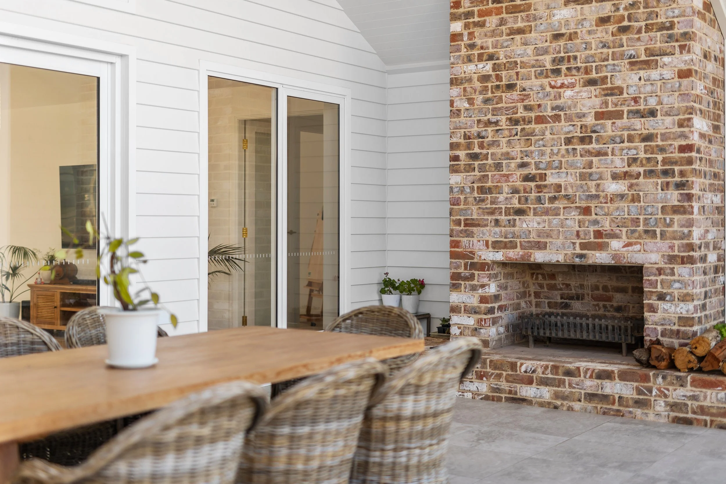Outdoor patio with wooden dining table, wicker chairs, potted plants, brick fireplace, and sliding glass doors leading indoors.