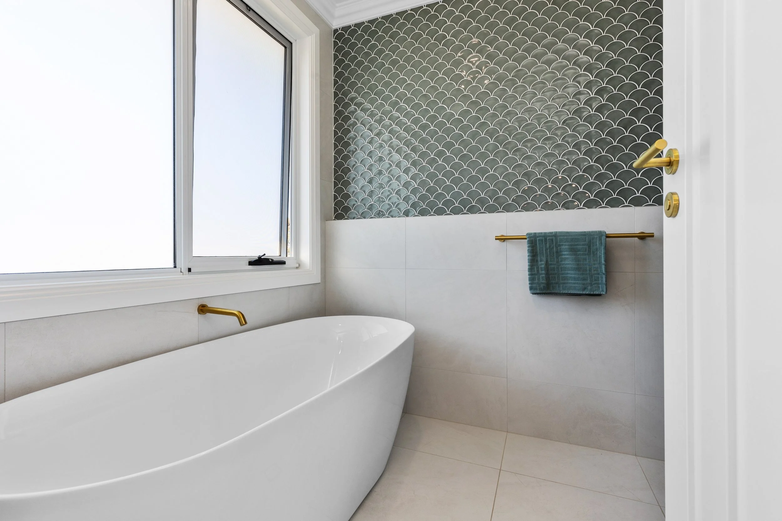 Modern bathroom with a white freestanding bathtub, gold fixtures, a frosted window, a teal towel, and decorative black scallop-patterned tiles on the wall.