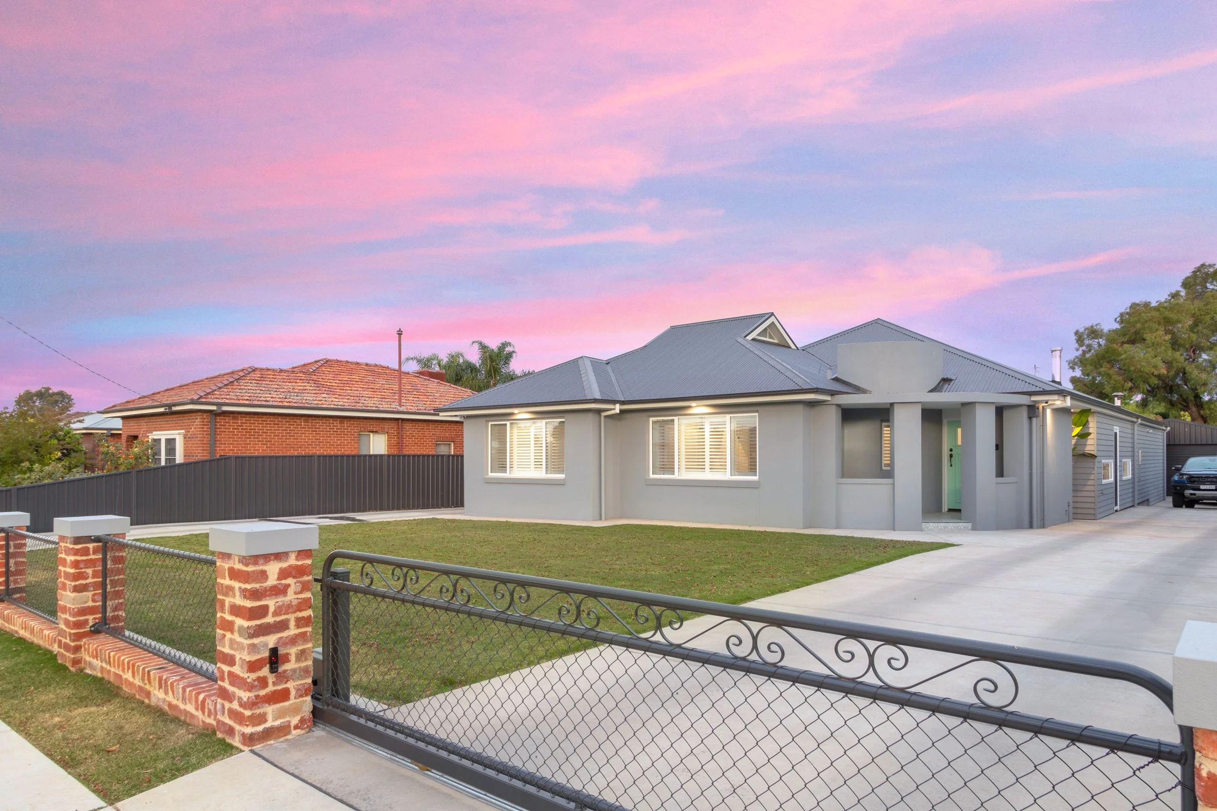 A modern, light gray house with a metal roof, a green front door, and a small front yard with a grass lawn. The house has large windows with white shutters and is surrounded by a black metal and brick fence. There is a driveway with a black car parked on it, and the sky above shows pink and purple hues during sunset.