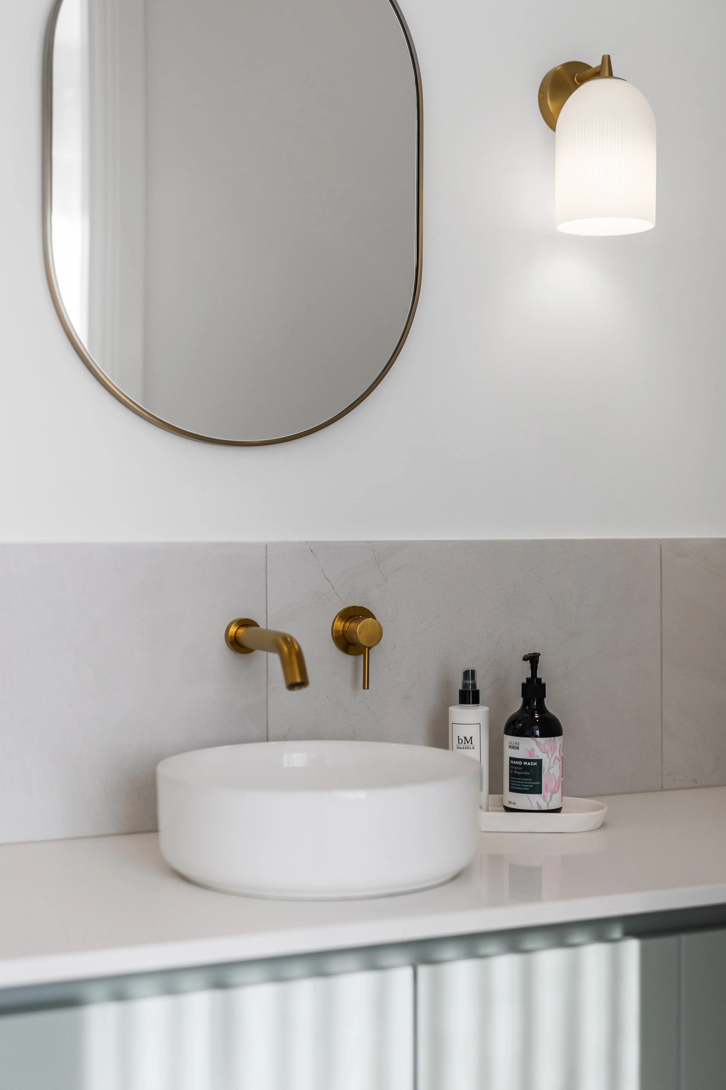 Modern bathroom sink with white countertop, oval mirror, brass faucet handles, and a wall-mounted light fixture with a white shade. Bottles of hand soap and lotion are on a tray beside the sink.