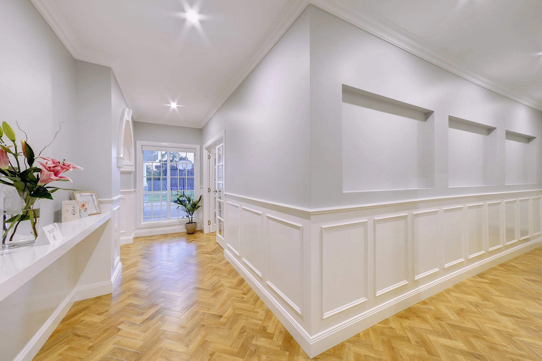Empty and bright hallway with white walls and wooden parquet flooring, decorated with a few framed pictures and flowers on a white shelf, leading to a window and a glass door.