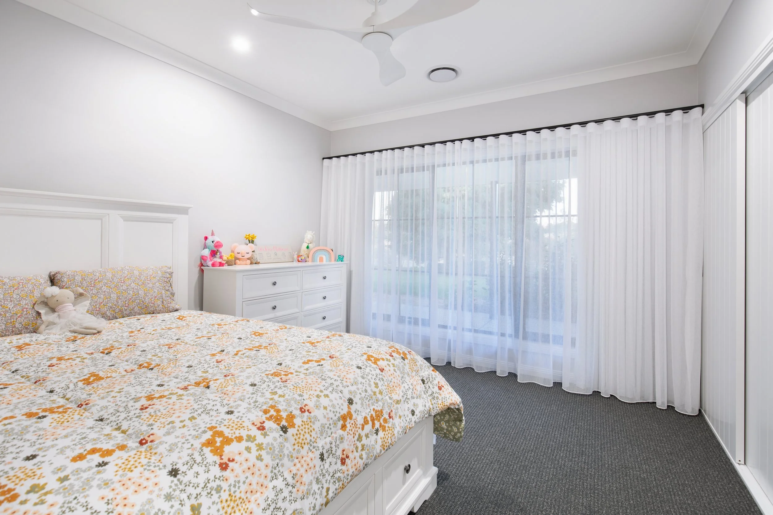 Bright child's bedroom with white walls, a bed with floral bedding, and a white dresser decorated with plush toys and colorful items. Large windows with sheer white curtains.