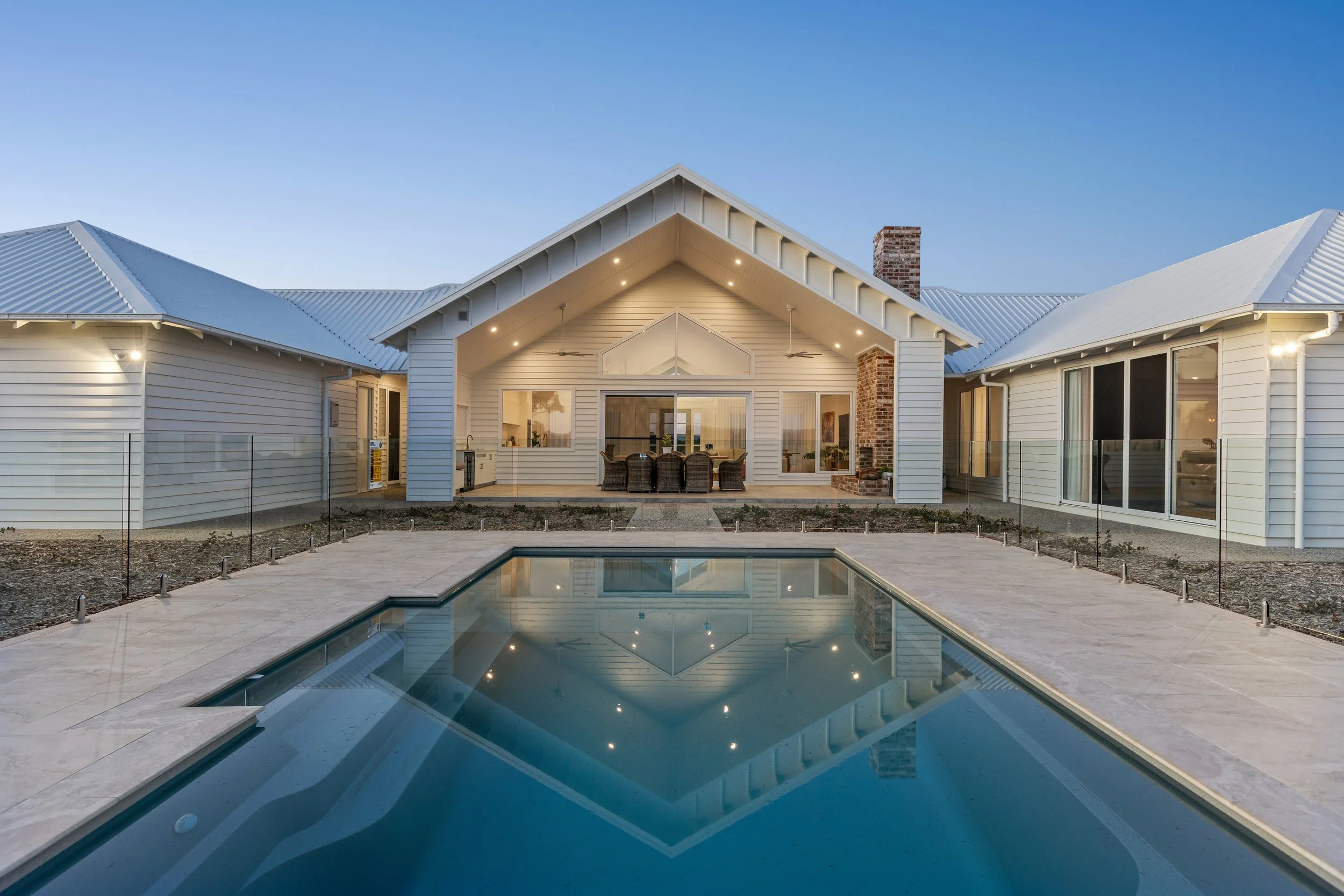 A modern house with a white roof, large glass windows, and a backyard swimming pool reflecting the house and blue sky.