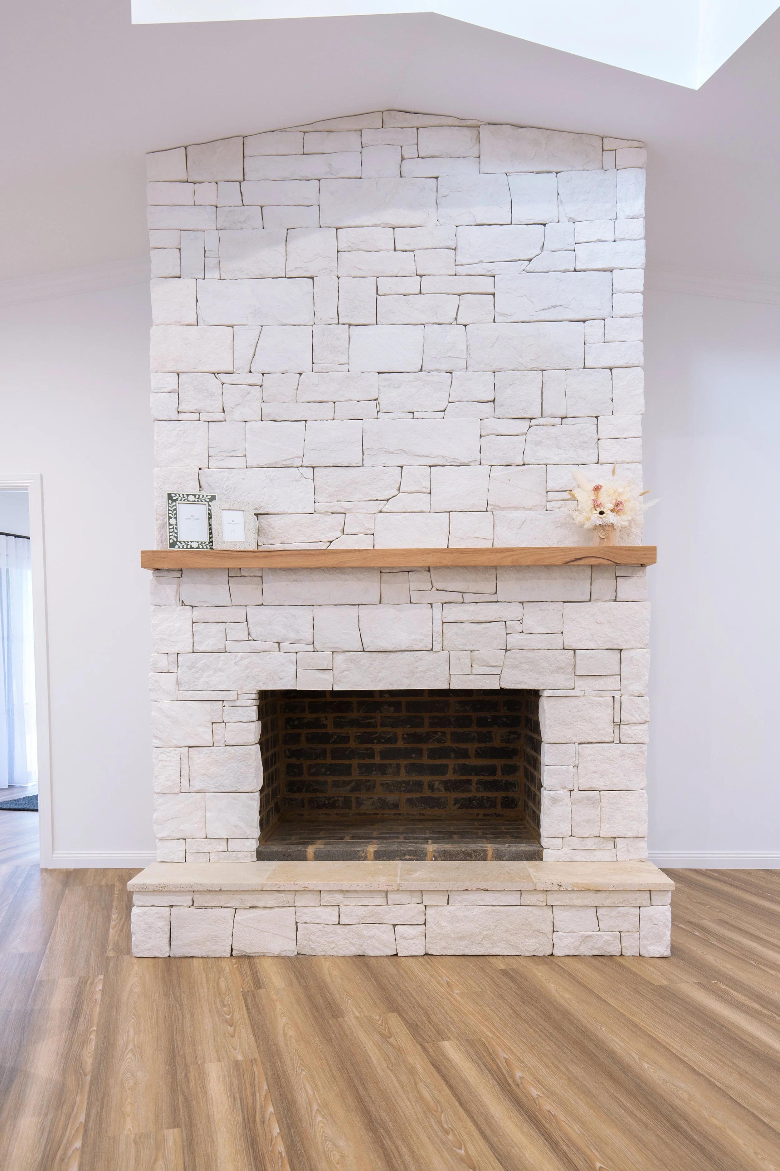 A white stone fireplace with a wooden mantel, decorated with picture frames and a floral arrangement, in a modern living room with wooden flooring.