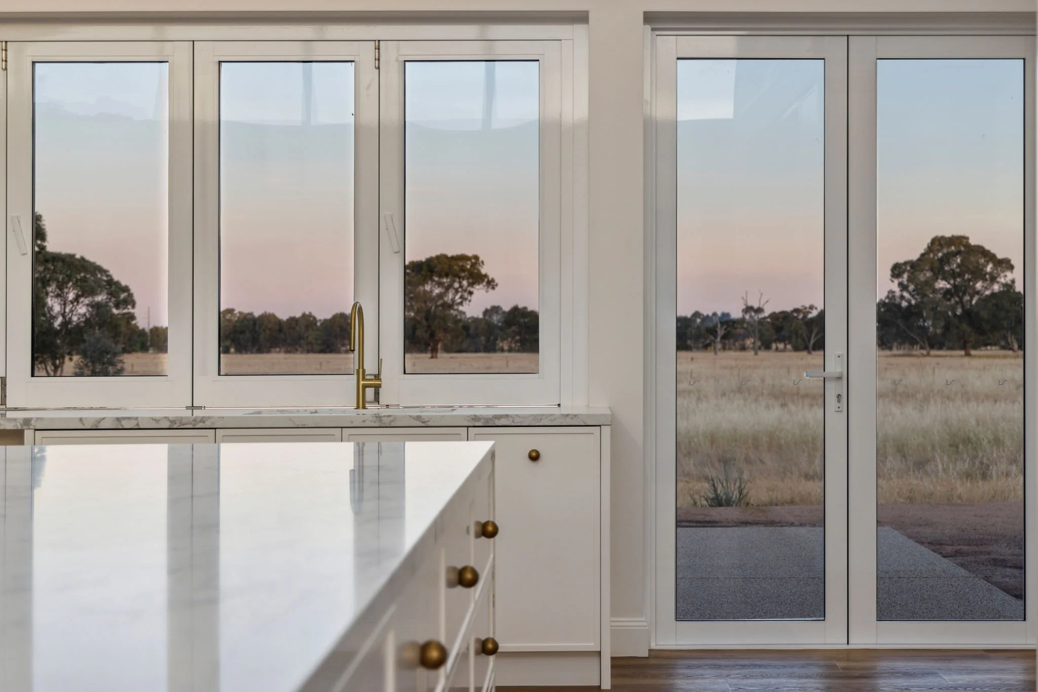 View of a kitchen with large windows and glass door, showing an outdoor field with grass and trees at sunset.