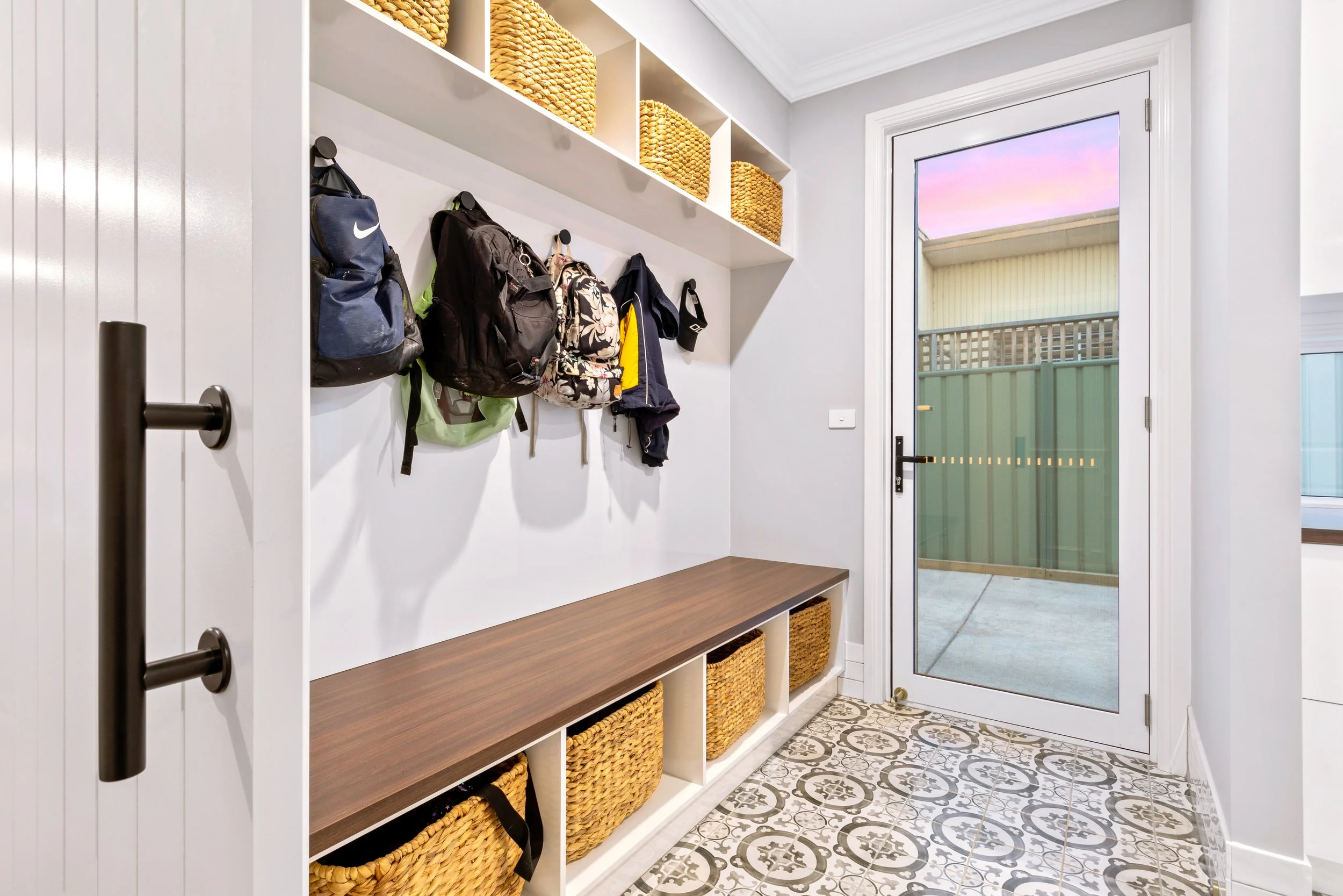 Entryway with white walls, a wooden bench with storage baskets underneath, hooks with backpacks and jackets, and shelves with woven baskets, next to a glass door leading outside.