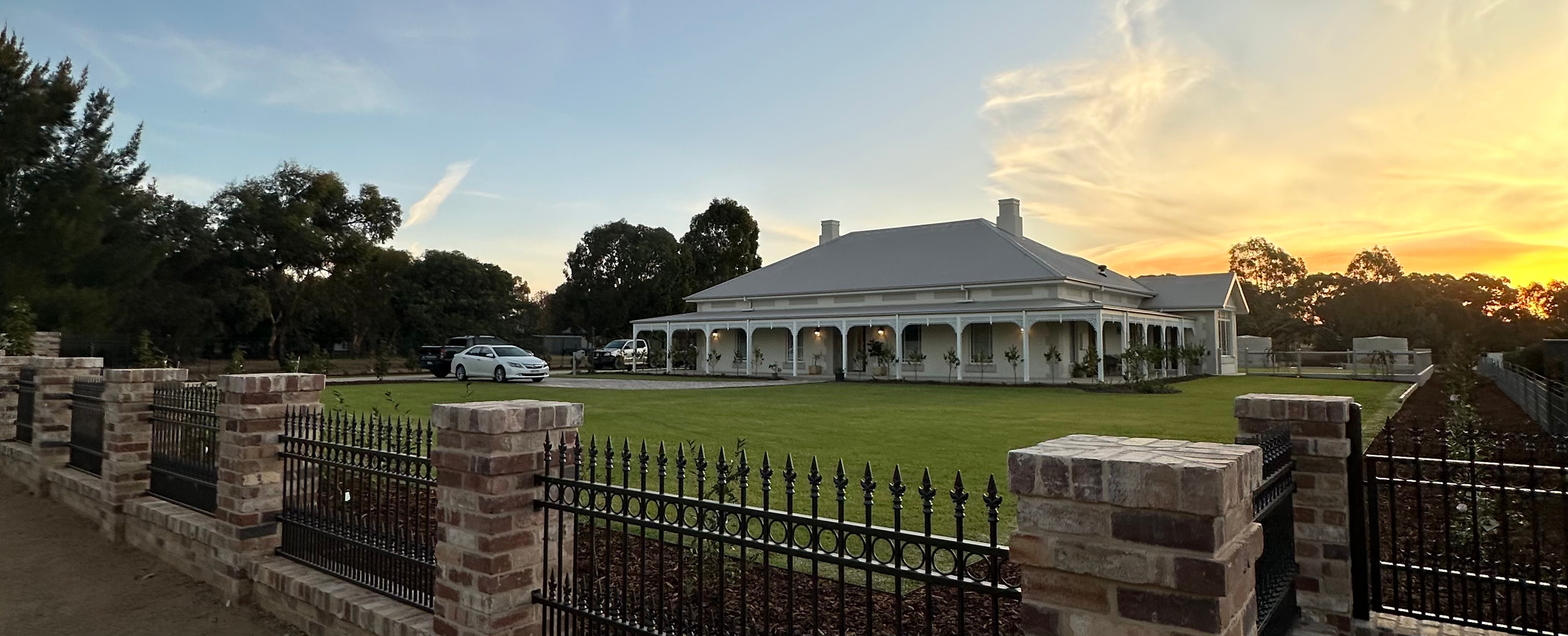 A large white house with a wraparound porch and multiple chimneys, surrounded by a well-maintained lawn and a brick and iron fence. Several cars are parked in front, and the sun is setting behind trees, casting a warm glow.