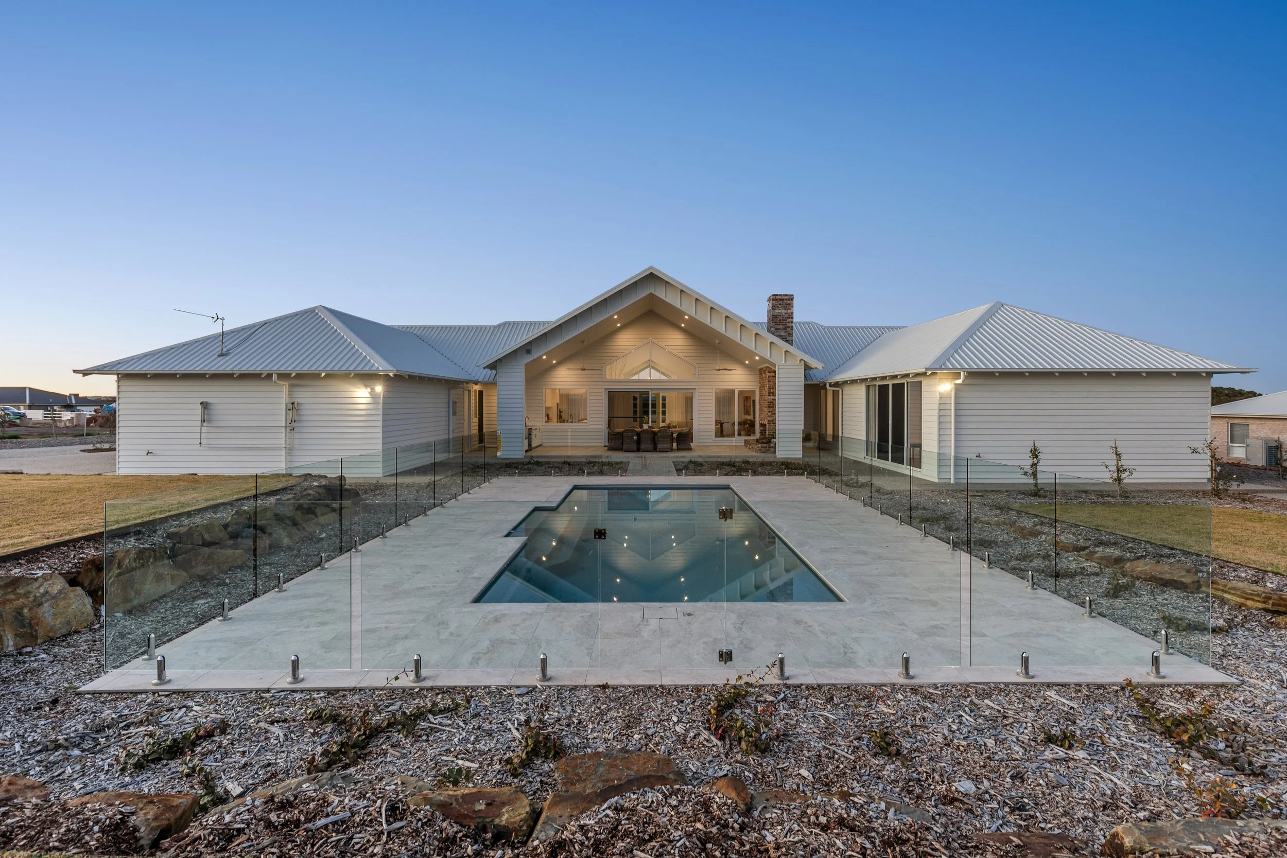 Modern house with white exterior, gray metal roof, and a backyard with a rectangular swimming pool enclosed by glass fencing, during dusk.