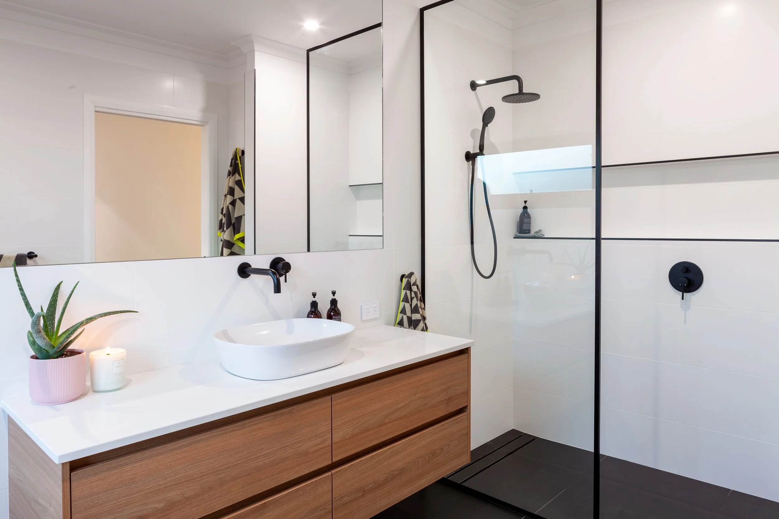 Modern bathroom with a wooden vanity, vessel sink, black faucet, large mirror, potted plant, candle, and a walk-in shower with black fixtures and a glass divider.