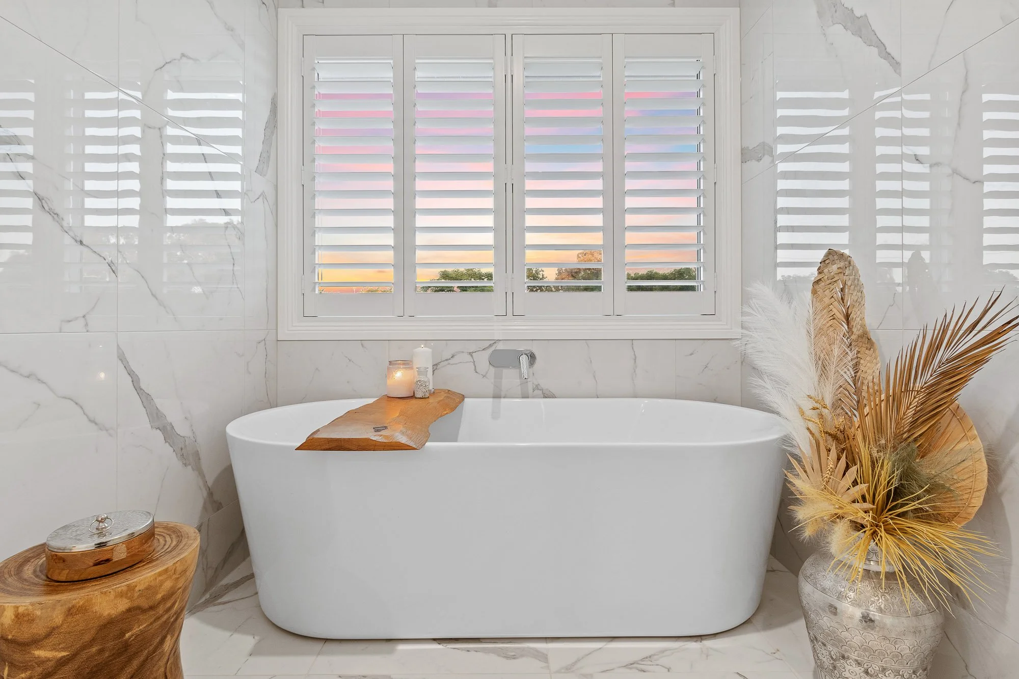 Modern bathroom with a white bathtub by a window with shutters, decorated with a wooden tray, candles, and a vase with dried plants.
