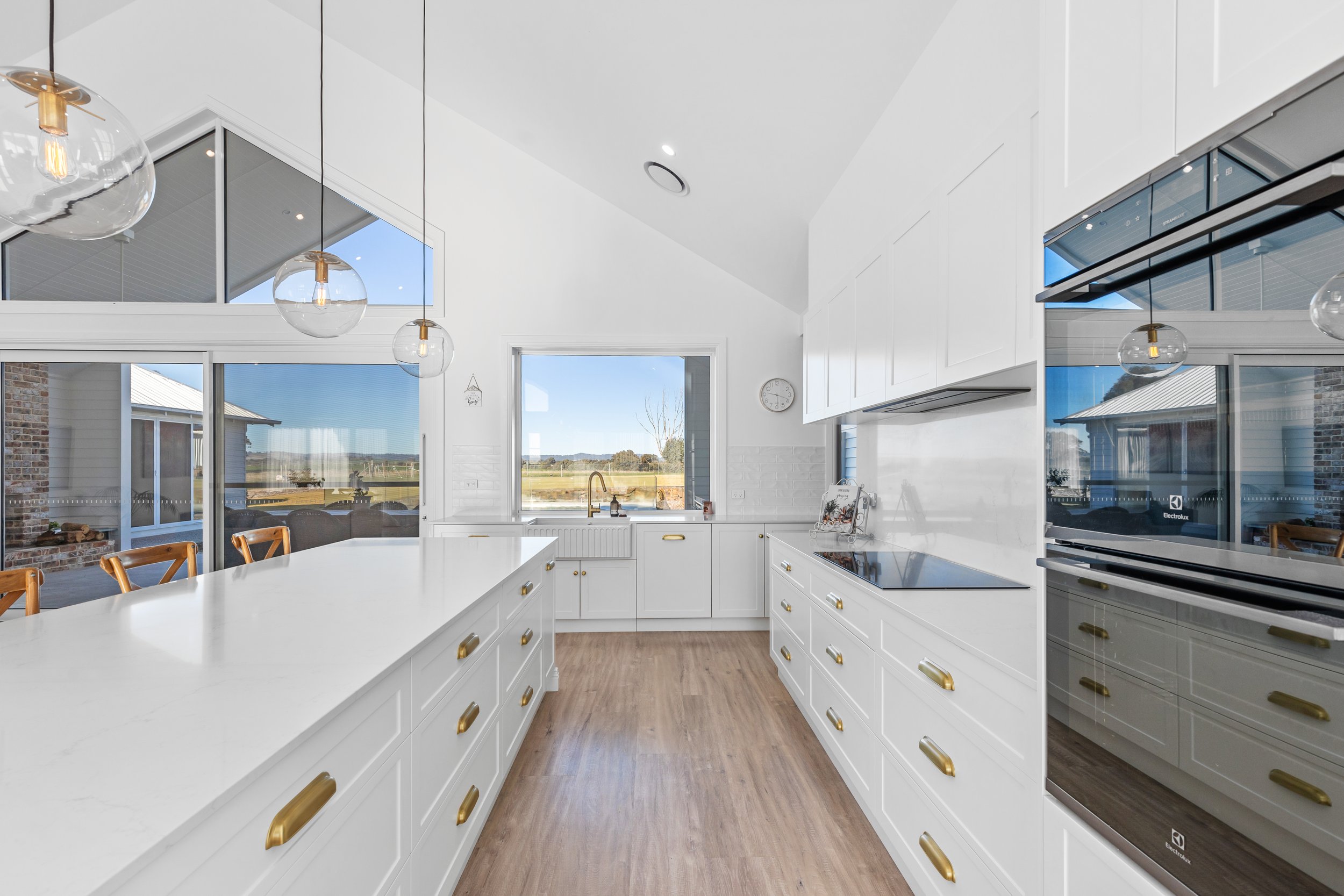 Modern white kitchen with gold hardware, large island, microwave, oven, windows showing outdoor landscape, pendant lights, and wooden flooring.