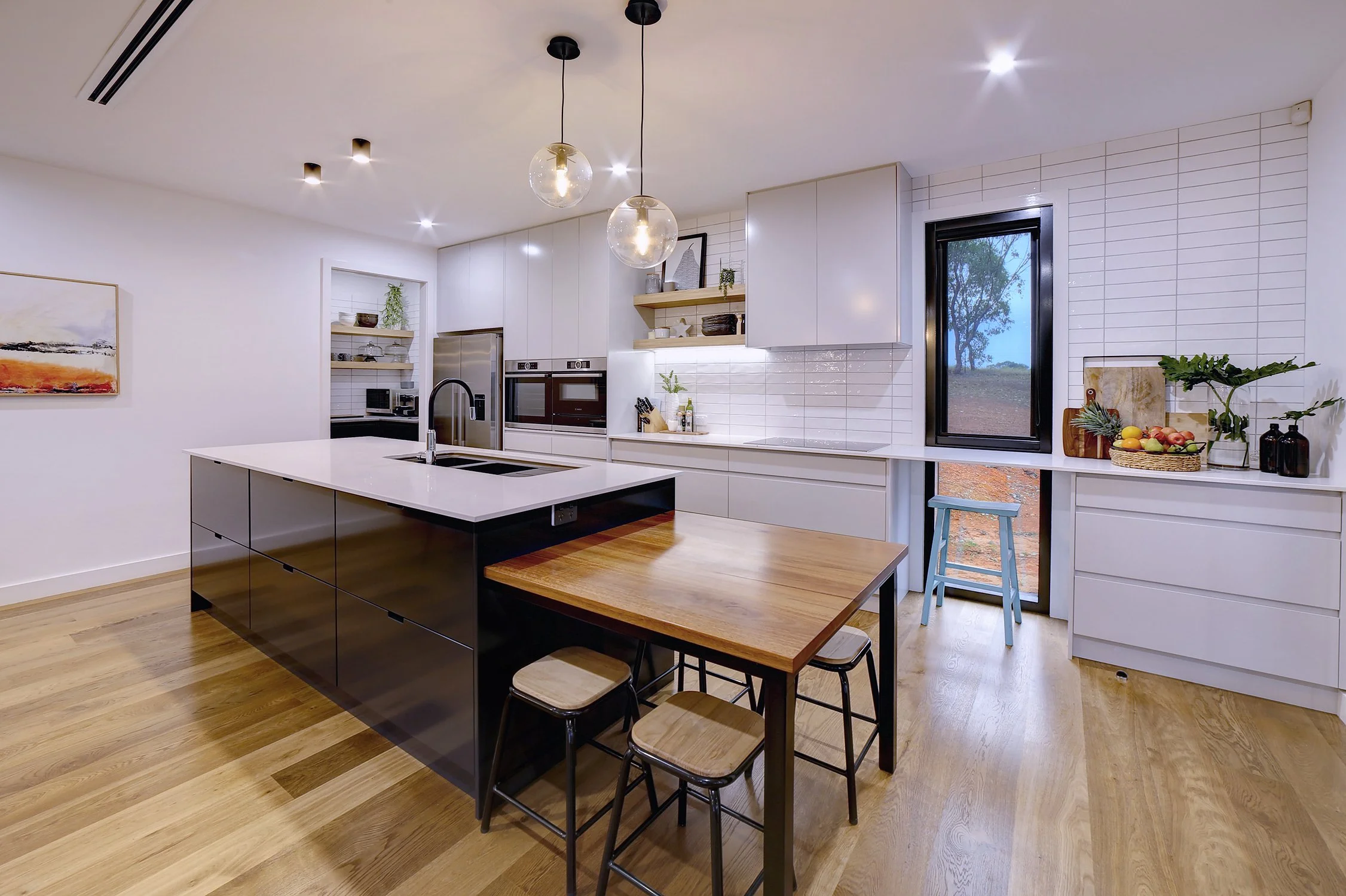 Modern kitchen with black island, white cabinets, wooden dining table, fruit basket, and window overlooking outdoor landscape.