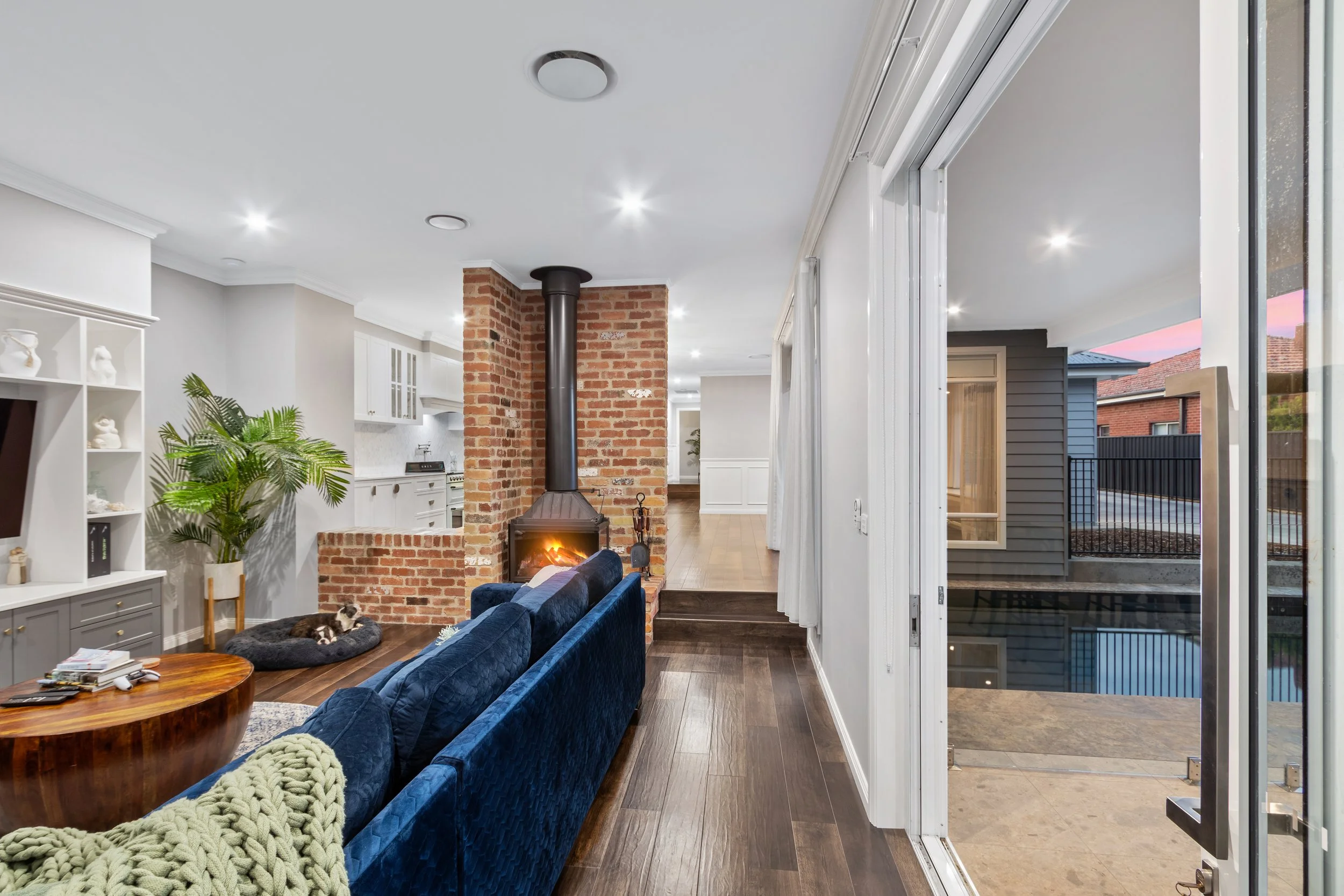 Living room with a brick fireplace, navy blue velvet sofa, wood coffee table, white built-in shelves, and large sliding glass doors leading outside at sunset.