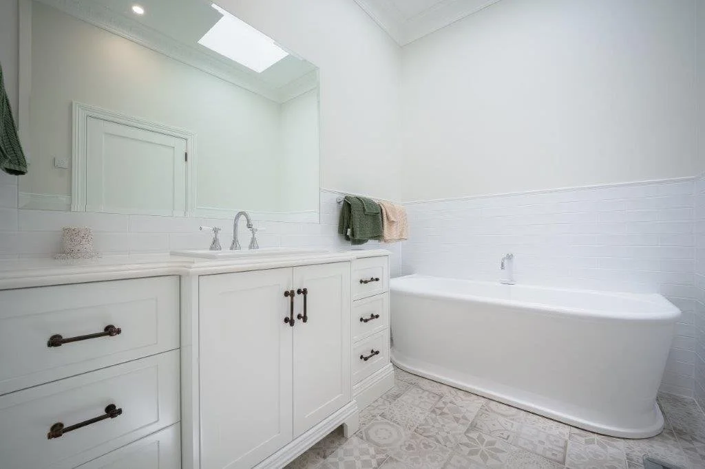 A bright, minimalist bathroom with a large wall mirror, white vanity with black handles, and a bathtub. The vanity has a sink and a small decorative object on the left side. Towels are hanging on the wall, and the room has a skylight and patterned tile flooring.