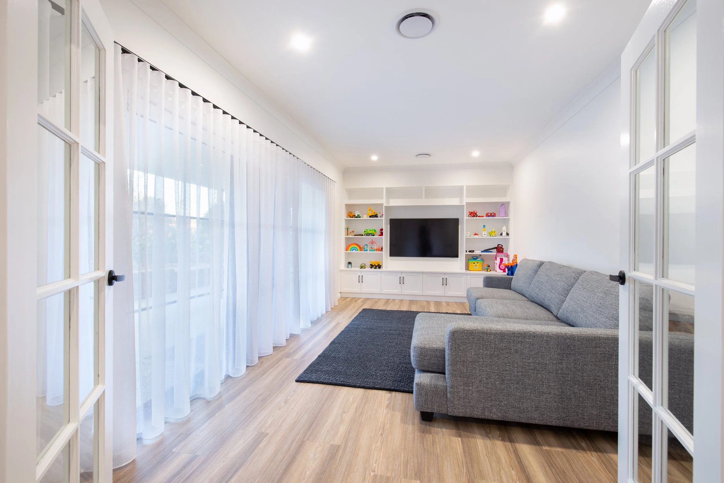 Living room with large window covered by white curtains, a gray sofa, a dark gray rug, and a built-in white entertainment center with toys and a flat-screen TV.