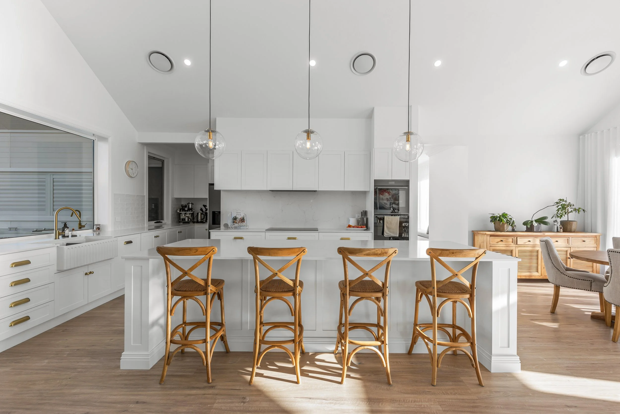 Modern white kitchen with a breakfast bar, four wooden barstools, and hanging pendant lights. Light wood flooring and a sideboard with potted plants in the background.