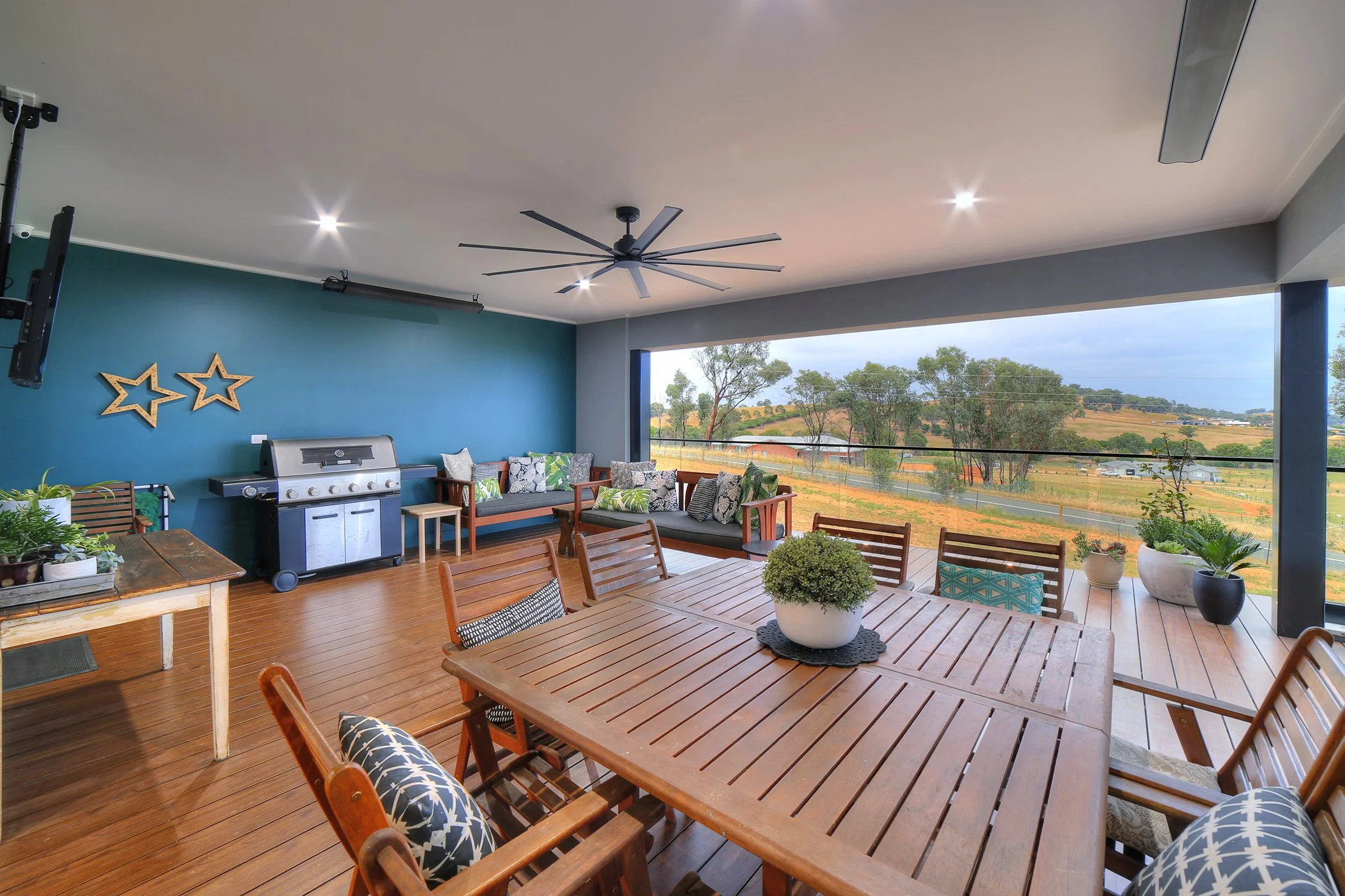 Outdoor patio with wooden dining table and chairs, potted plants, sofa with cushions, grill, and a large window revealing a landscape with trees and rolling hills.