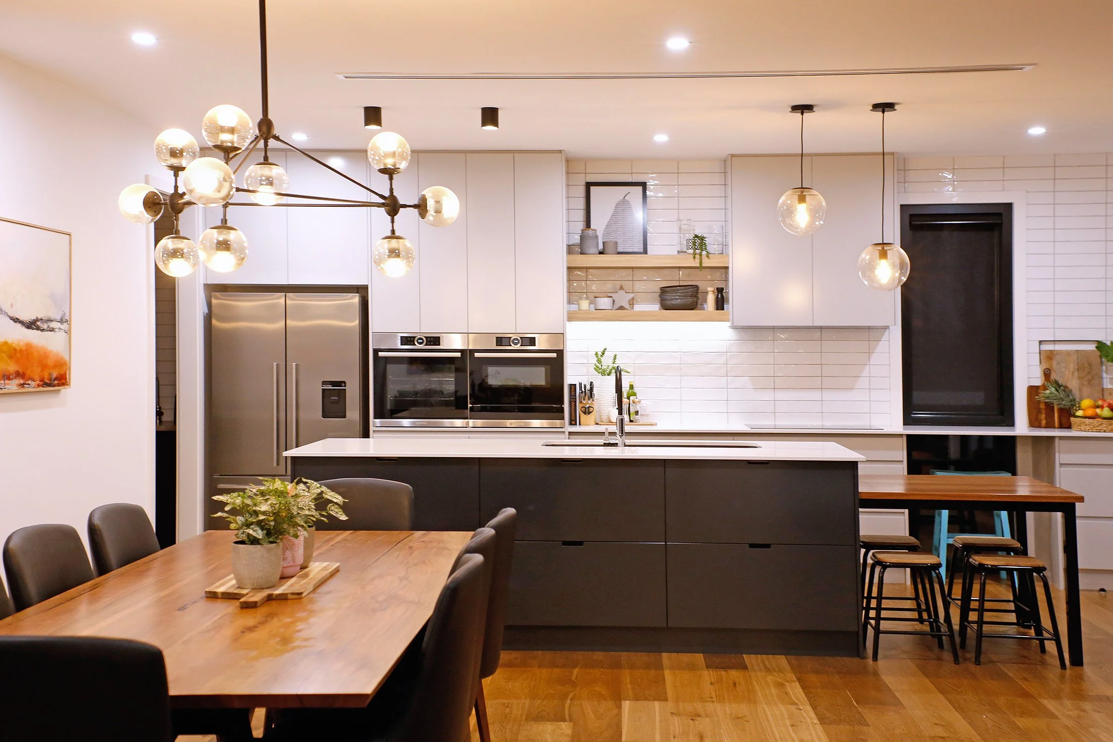 Modern kitchen with an island, open shelving, black and white cabinetry, pendant lighting, and a dining table with black chairs.