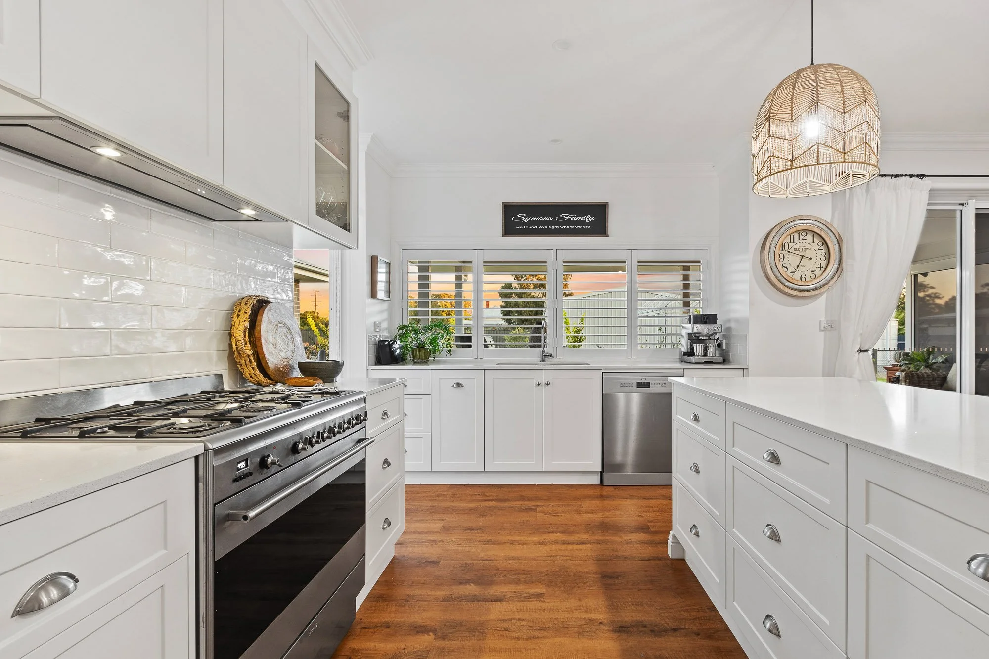 White kitchen with wooden flooring, stainless steel stove, white cabinets, and a window with shutters showing a sunset outside.