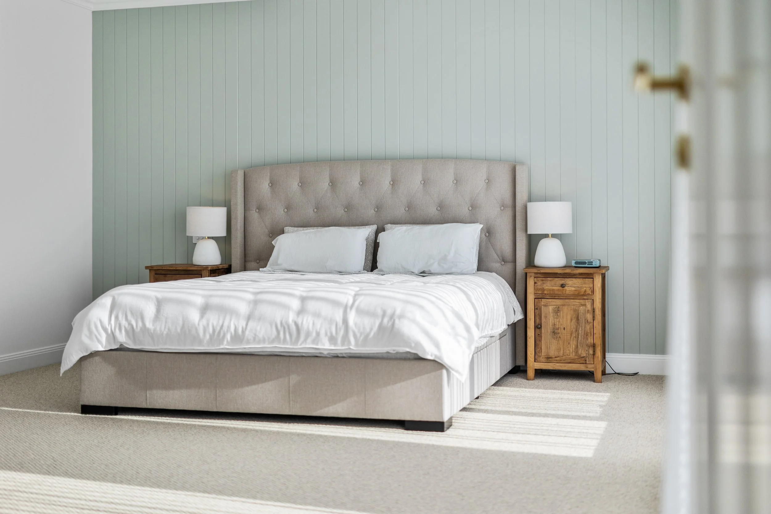 A bedroom with a neutral-colored bed, two matching nightstands with white lamps, light green wall, and white bedding.
