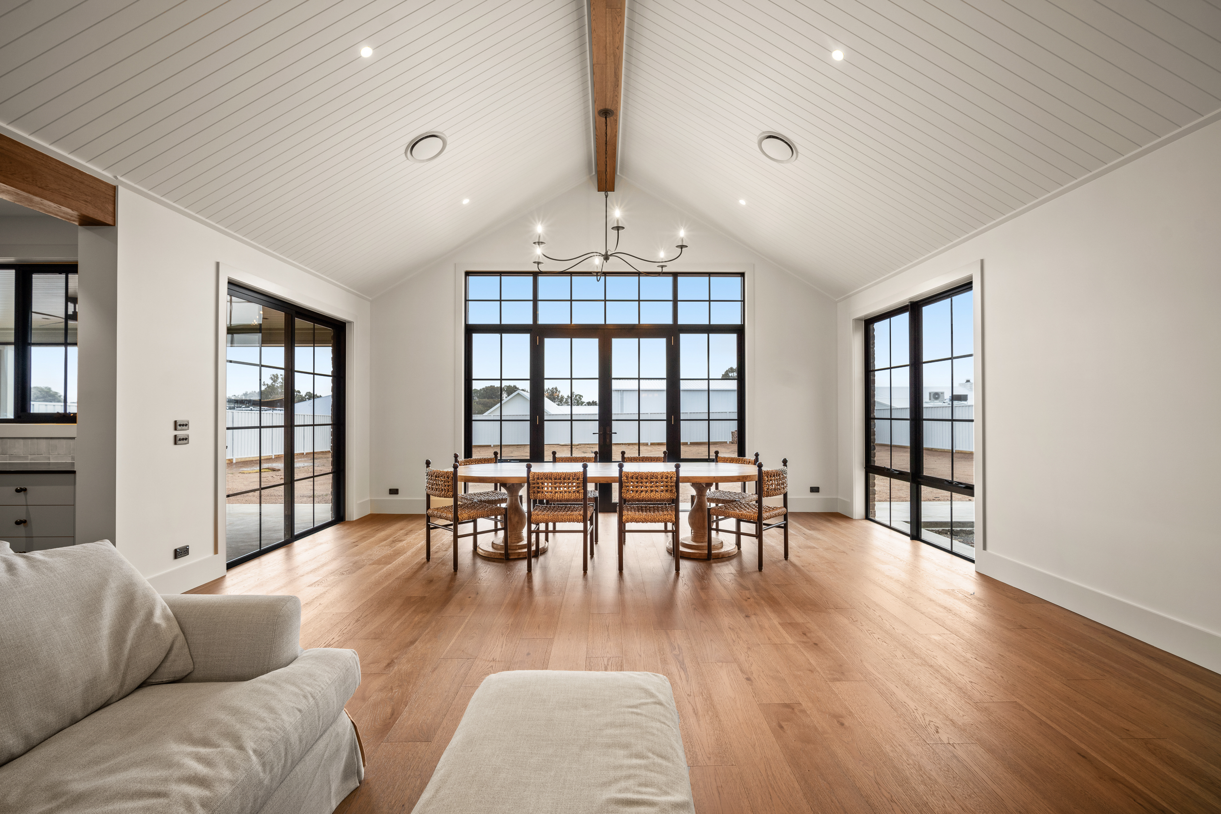 Bright modern dining room with a farmhouse table, woven chairs, vaulted white-paneled ceiling, chandelier, hardwood floors, and expansive black-framed windows.