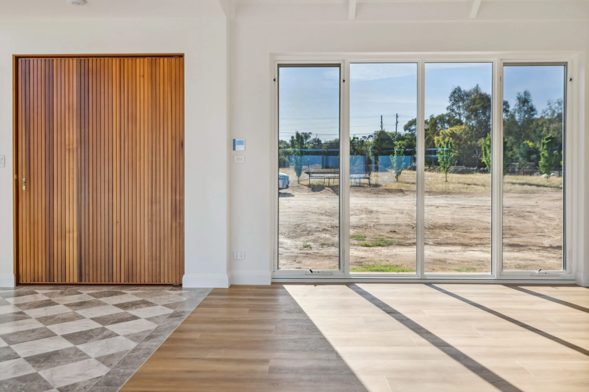 Interior view of a room with a sliding glass door leading outside, showing an outdoor area with trees, a bench, and a dirt ground. The room has a mix of hardwood and tiled flooring, white walls, and a wooden wall panel.