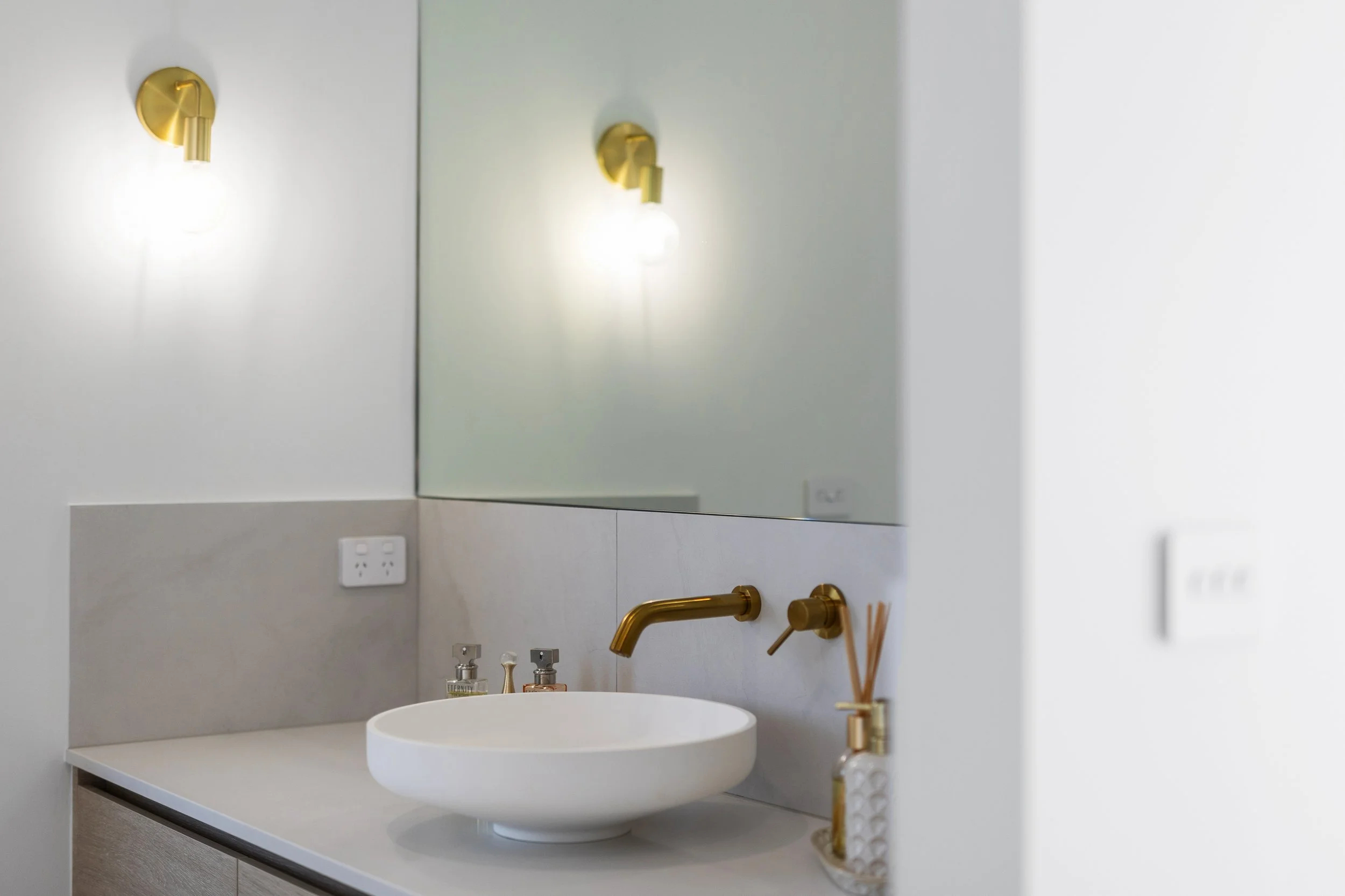 Modern bathroom vanity with a white vessel sink, gold faucet and fixtures, a large mirror, and a decorative reed diffuser on the countertop.