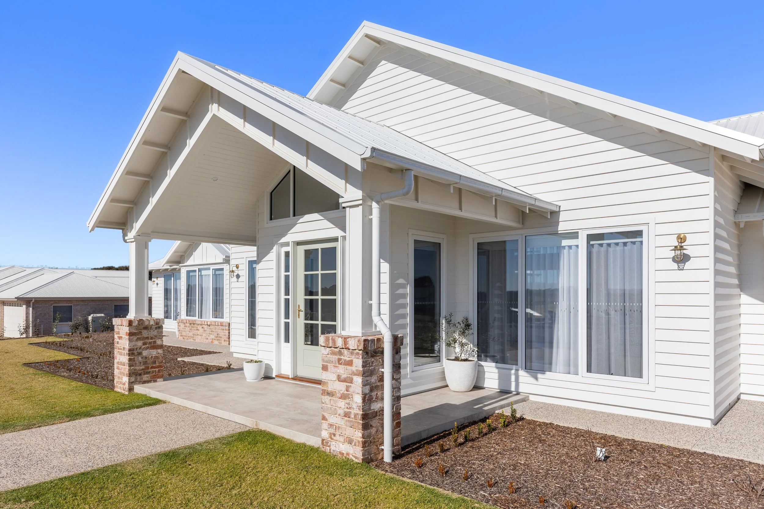 Exterior view of a modern white house with large windows, a small porch, brick accents, and a well-maintained lawn under a clear blue sky.