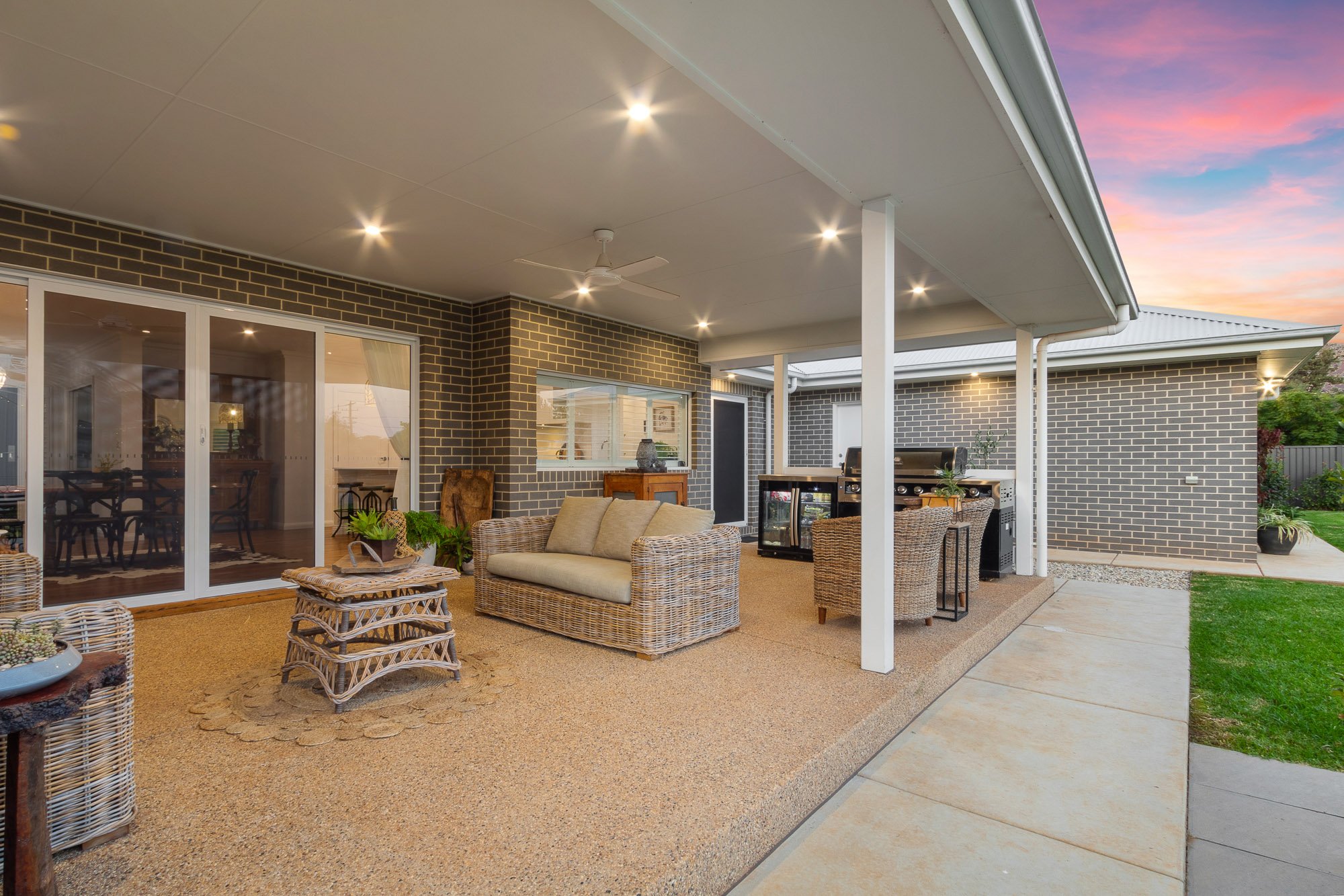 Covered outdoor patio with wicker furniture, including a sofa and chairs, an outdoor grill, and decorative plants, with a brick house exterior during sunset.