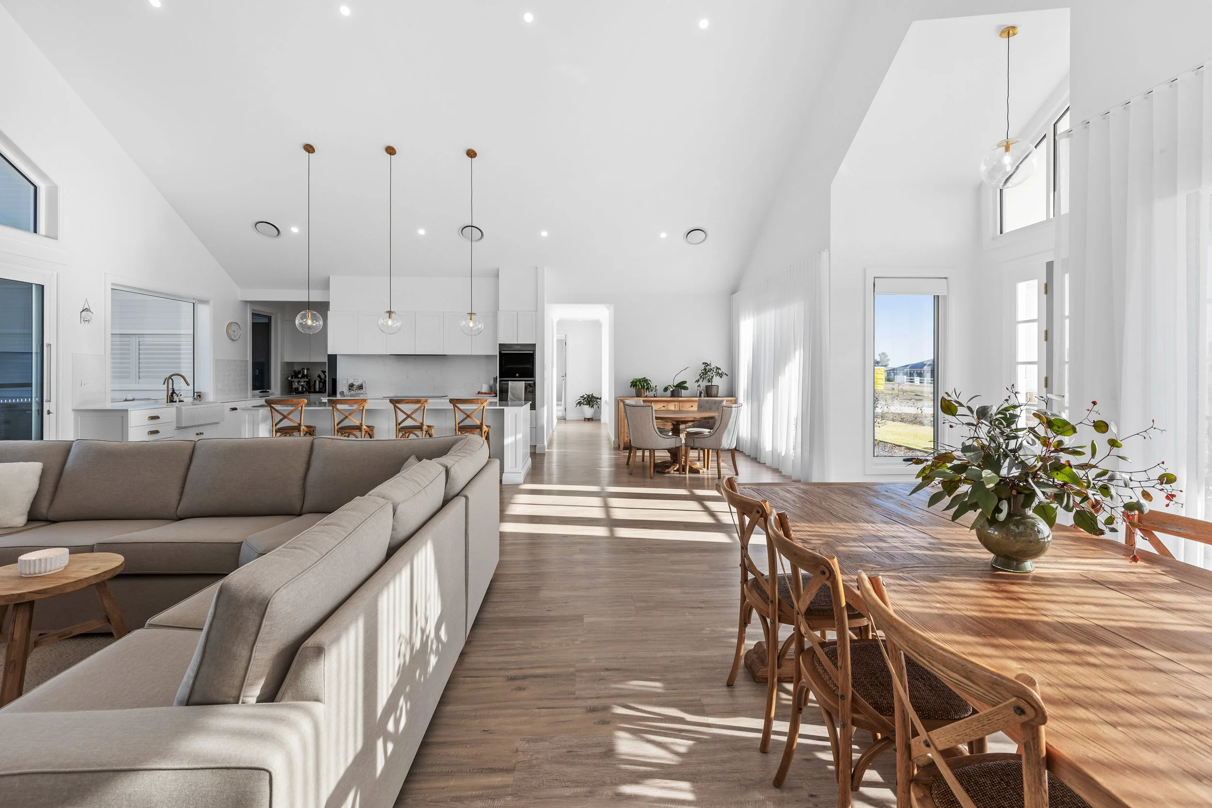 Open-concept living room and kitchen with white walls, large windows, and wooden furniture, including a dining table with a vase of flowers and a beige sofa.
